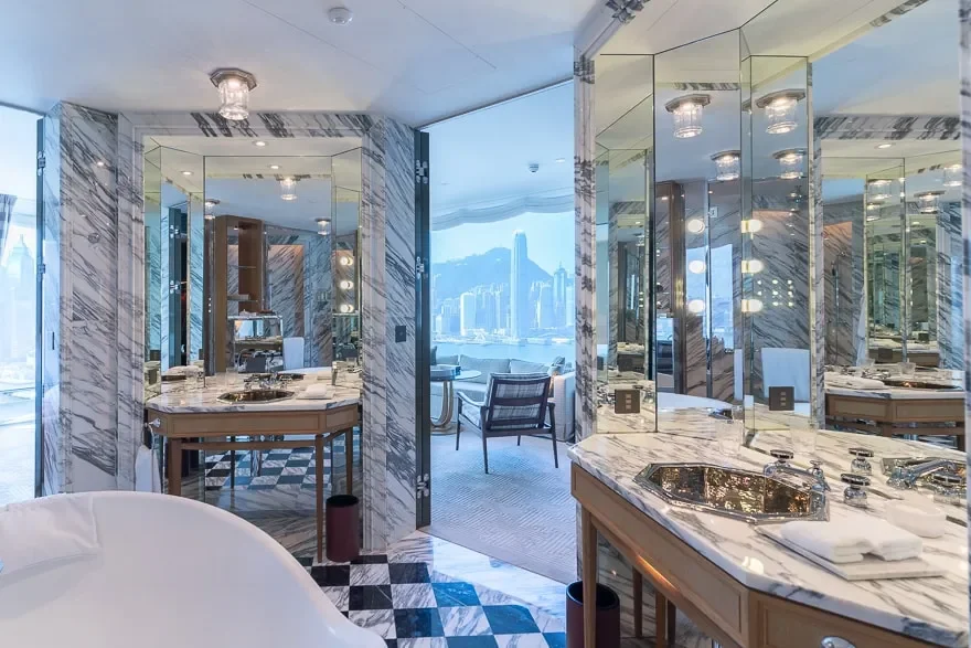Luxurious bathroom with marble walls and countertops, multiple mirrors with round light fixtures, a sink with toiletries, and a view of a city skyline and water through the windows.