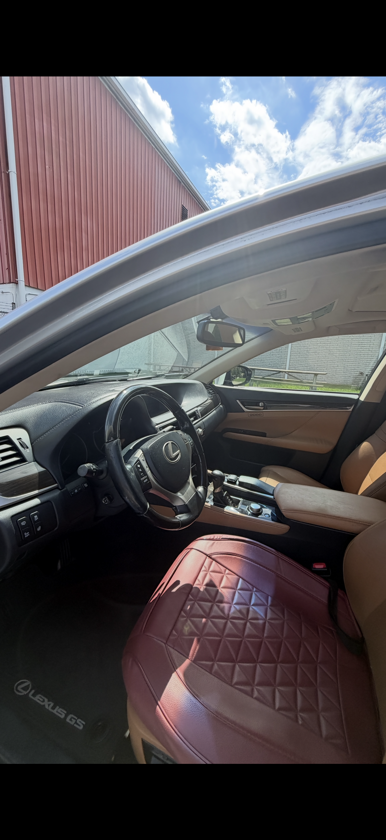 Interior of a Lexus GS vehicle with black dashboard, leather seats, maroon quilted seat covers, and a view of the exterior blue sky and red building through the open window.