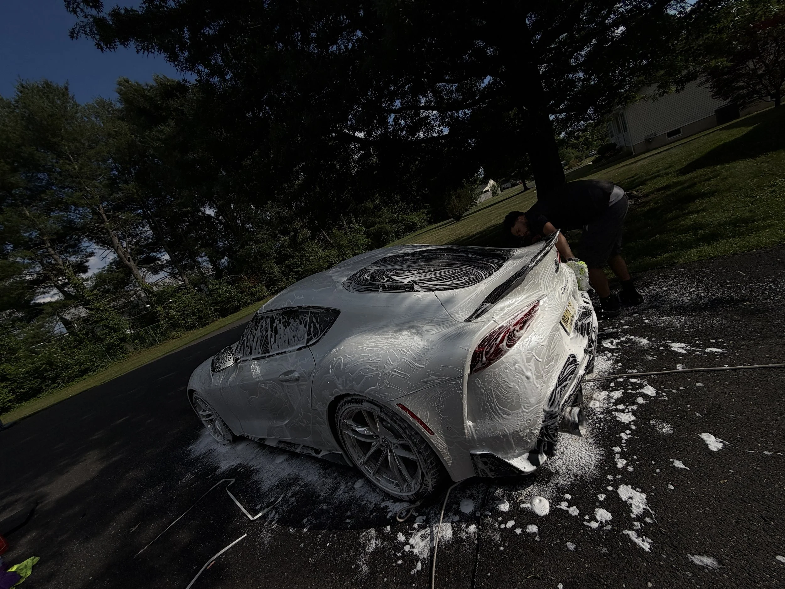 A person washing a white luxury sports car with soap and foam in a driveway, surrounded by trees and residential houses.