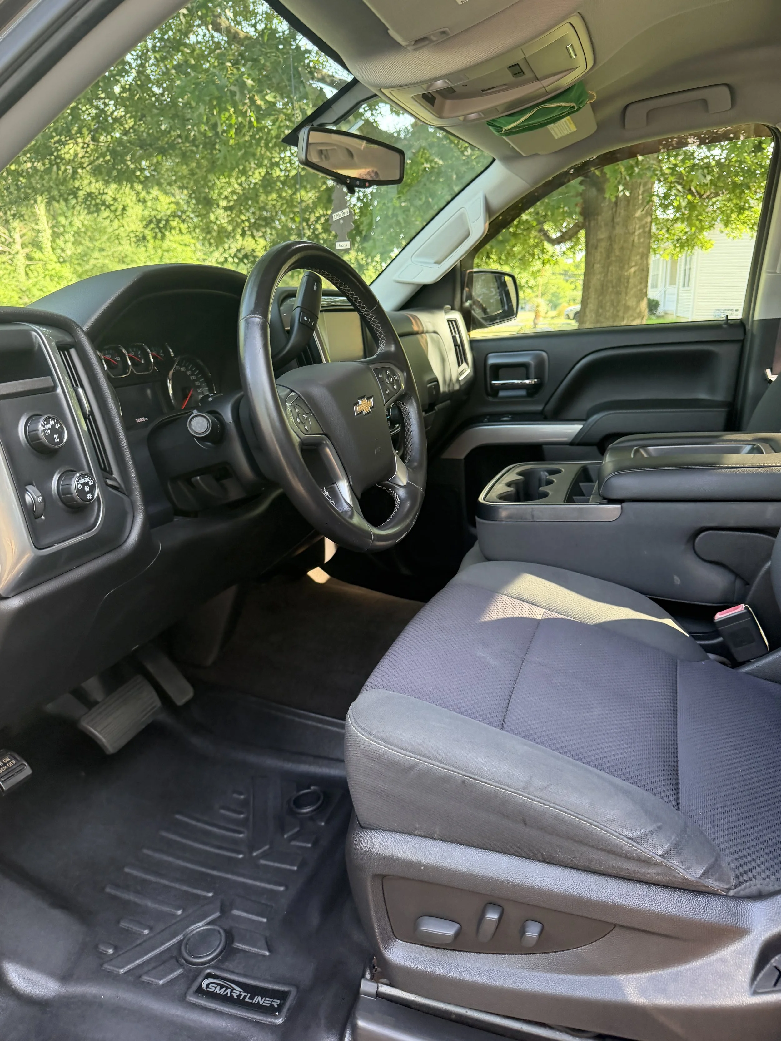 Interior of a Chevrolet vehicle showing the steering wheel, dashboard, driver's seat, center console, and floor mats, with a view of green trees outside through the windshield.