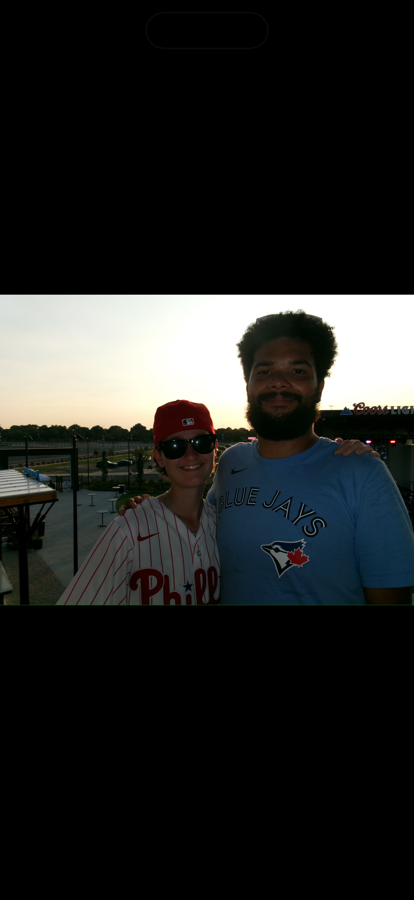 Two people standing outdoors at sunset, smiling, with a parking lot, trees, and a football stadium in the background. One person is wearing a Philadelphia Phillies baseball jersey, a red cap, and sunglasses, and the other is wearing a Toronto Blue Jays T-shirt.