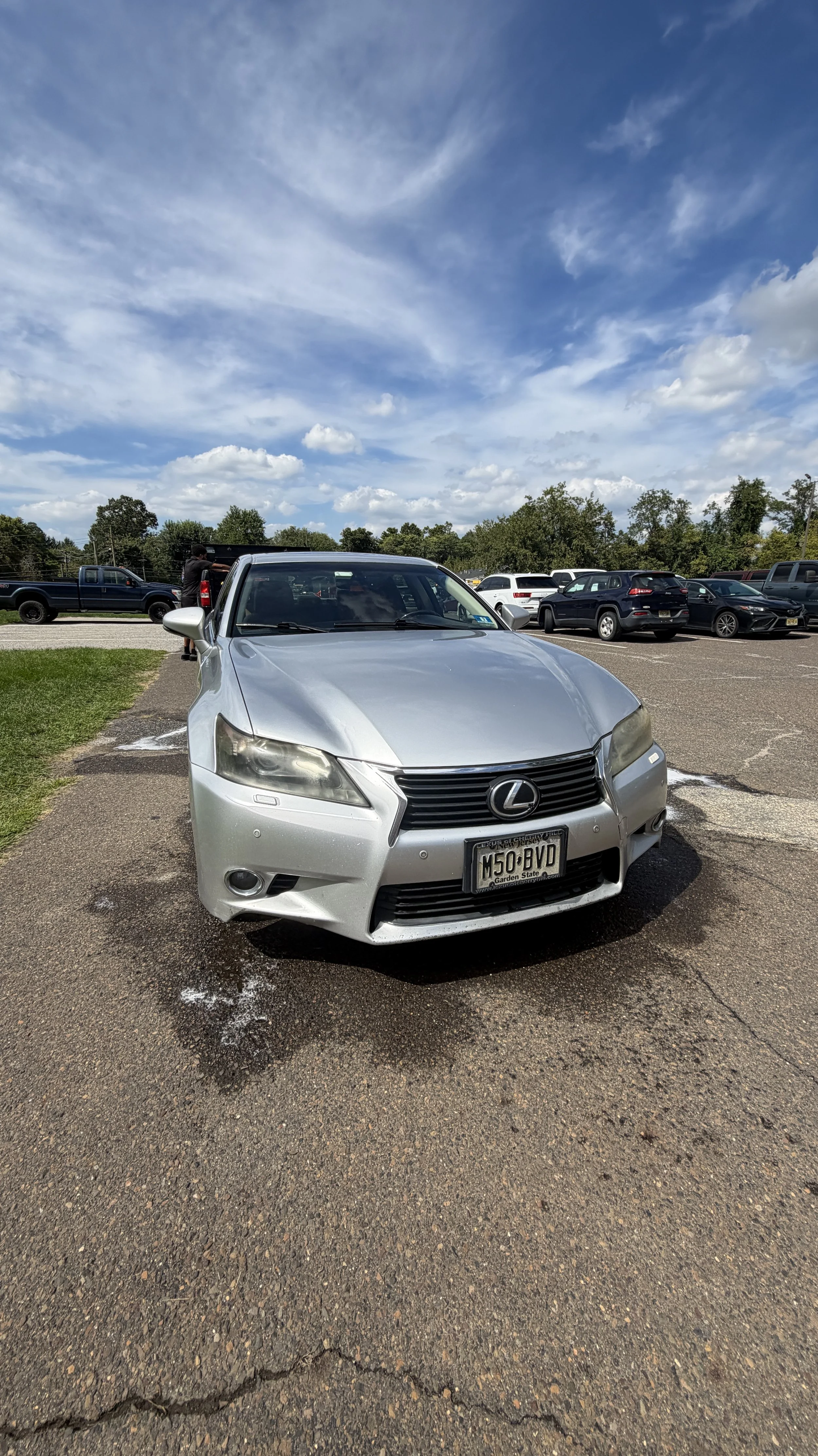 A silver Lexus car parked in a parking lot with a blue sky and clouds above, and multiple other cars visible in the background.