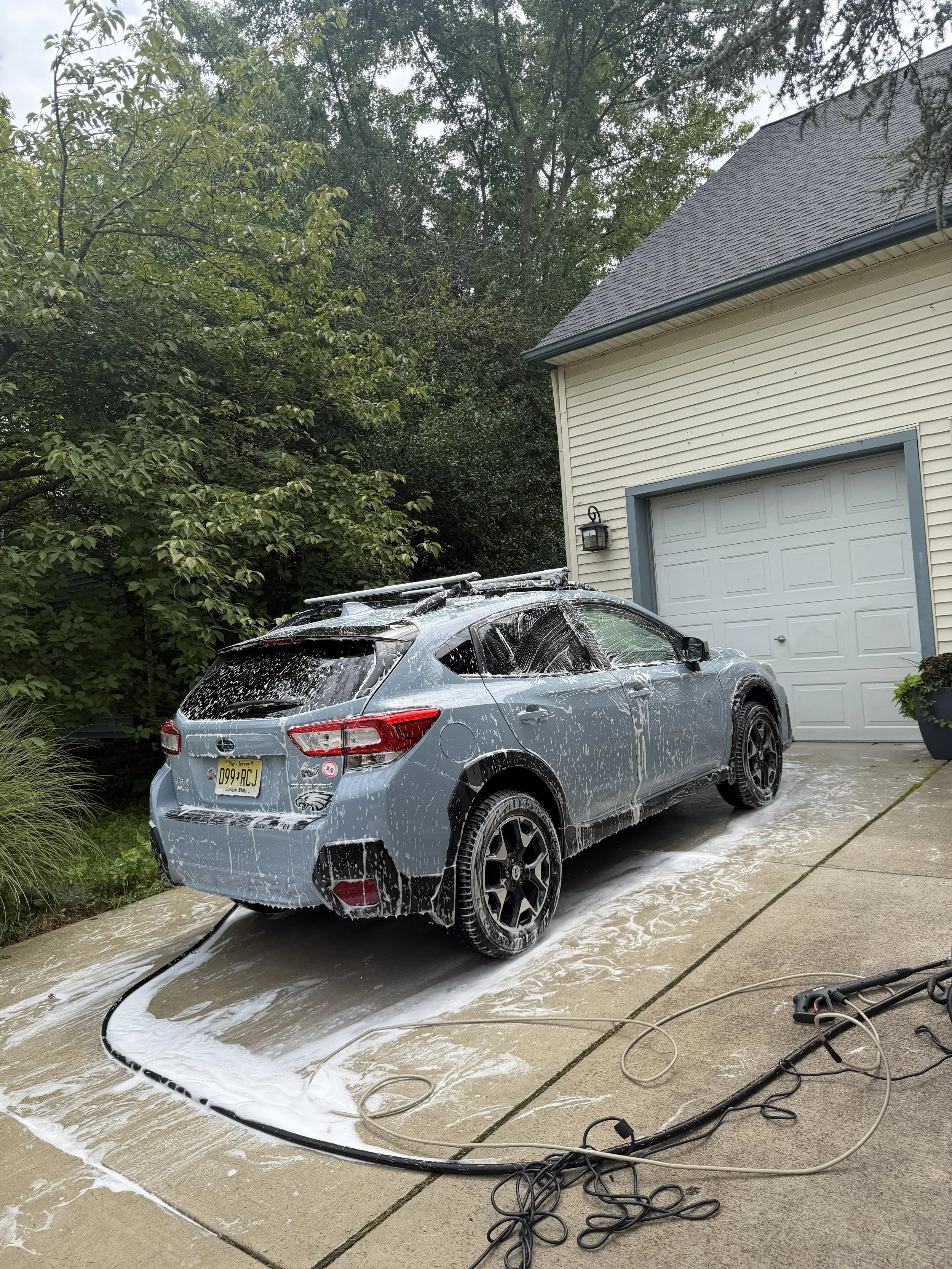 A silver compact SUV covered in soap suds is being washed on a concrete driveway. The car has a roof rack and a New Jersey license plate. A green plant is on the left side, and a white garage with a closed door is on the right side, with trees and bushes in the background. A pressure washer and hose are on the ground.