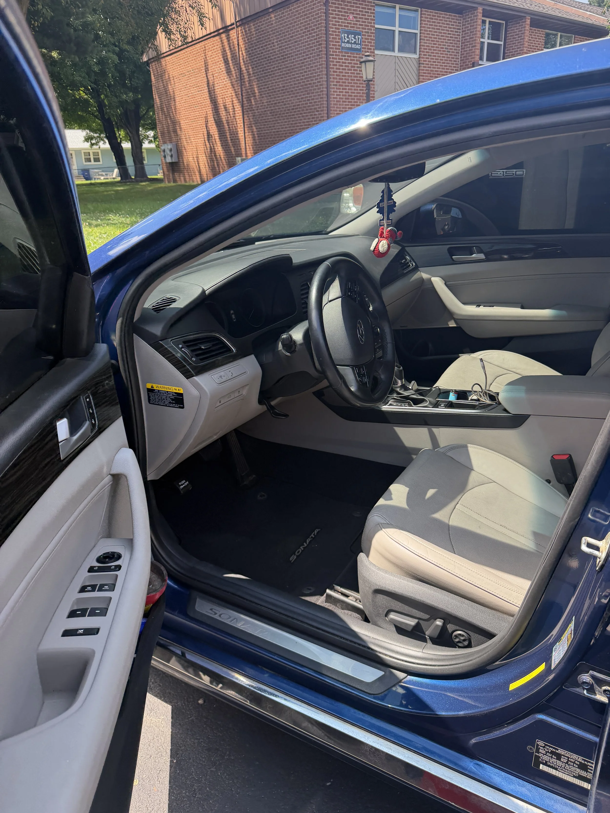 The interior of a blue Hyundai Sonata with the driver’s side door open, showing the front seats, dashboard, steering wheel, and hanging decorations. The background features a residential area with red brick houses and trees.