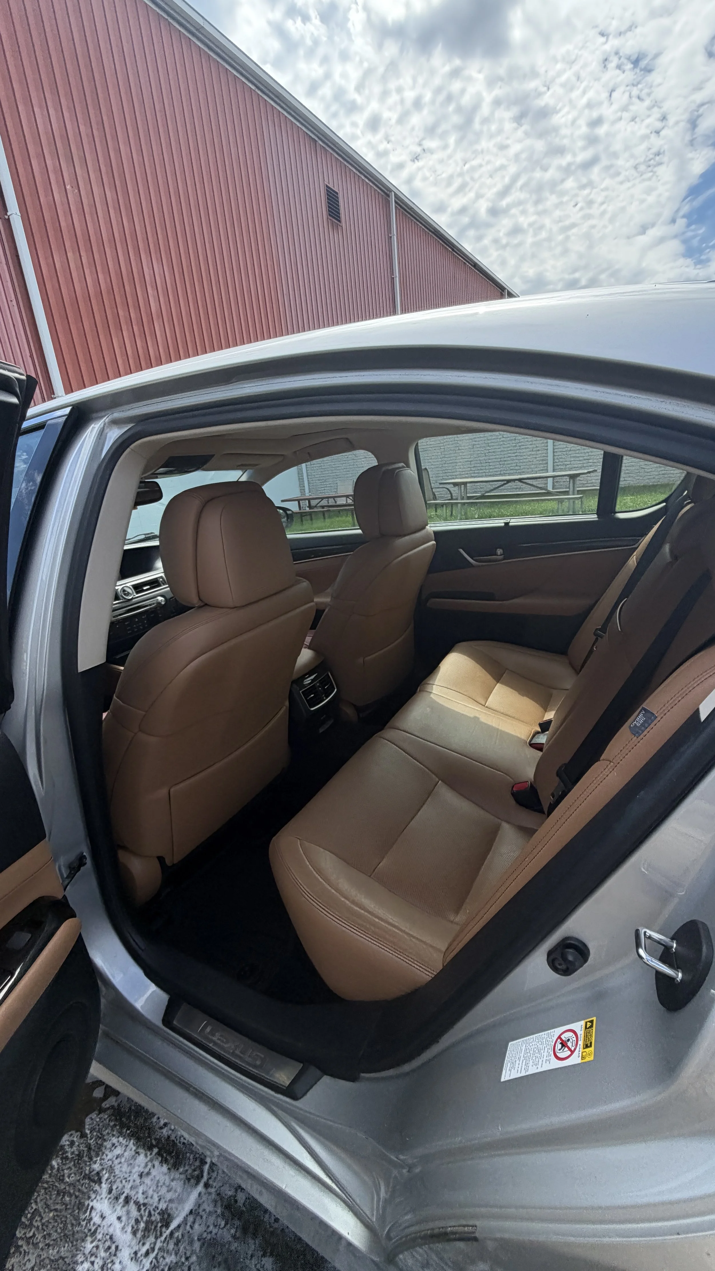View of the interior of a car with tan leather seats, showing the back seats and the front headrests. The window reveals an outdoor setting with a red building and a partly cloudy sky.