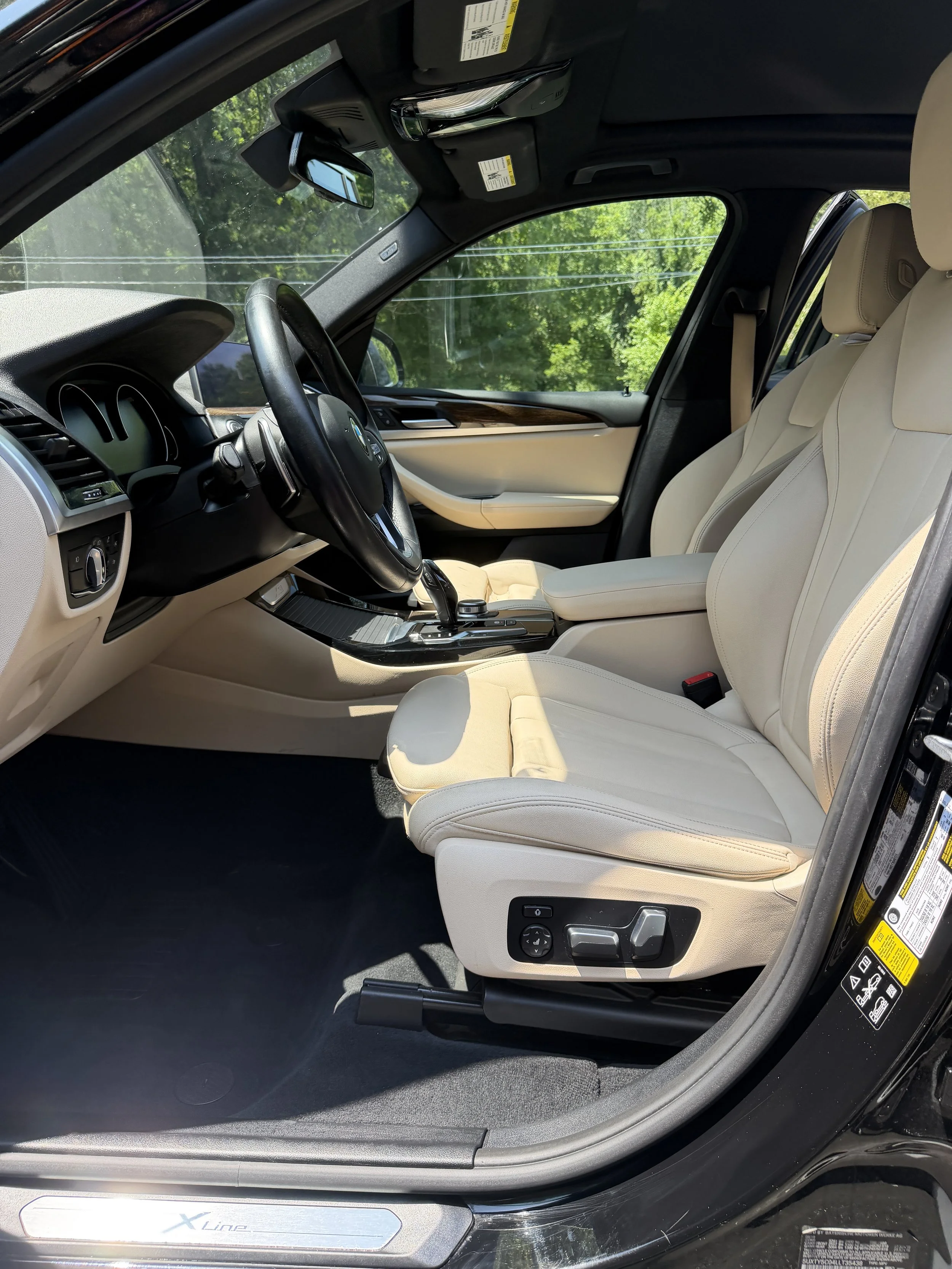 The interior of a luxury vehicle with beige leather seats, black dashboard, and a gear shift in the center console, viewed from the driver's side door open, with green trees visible outside.