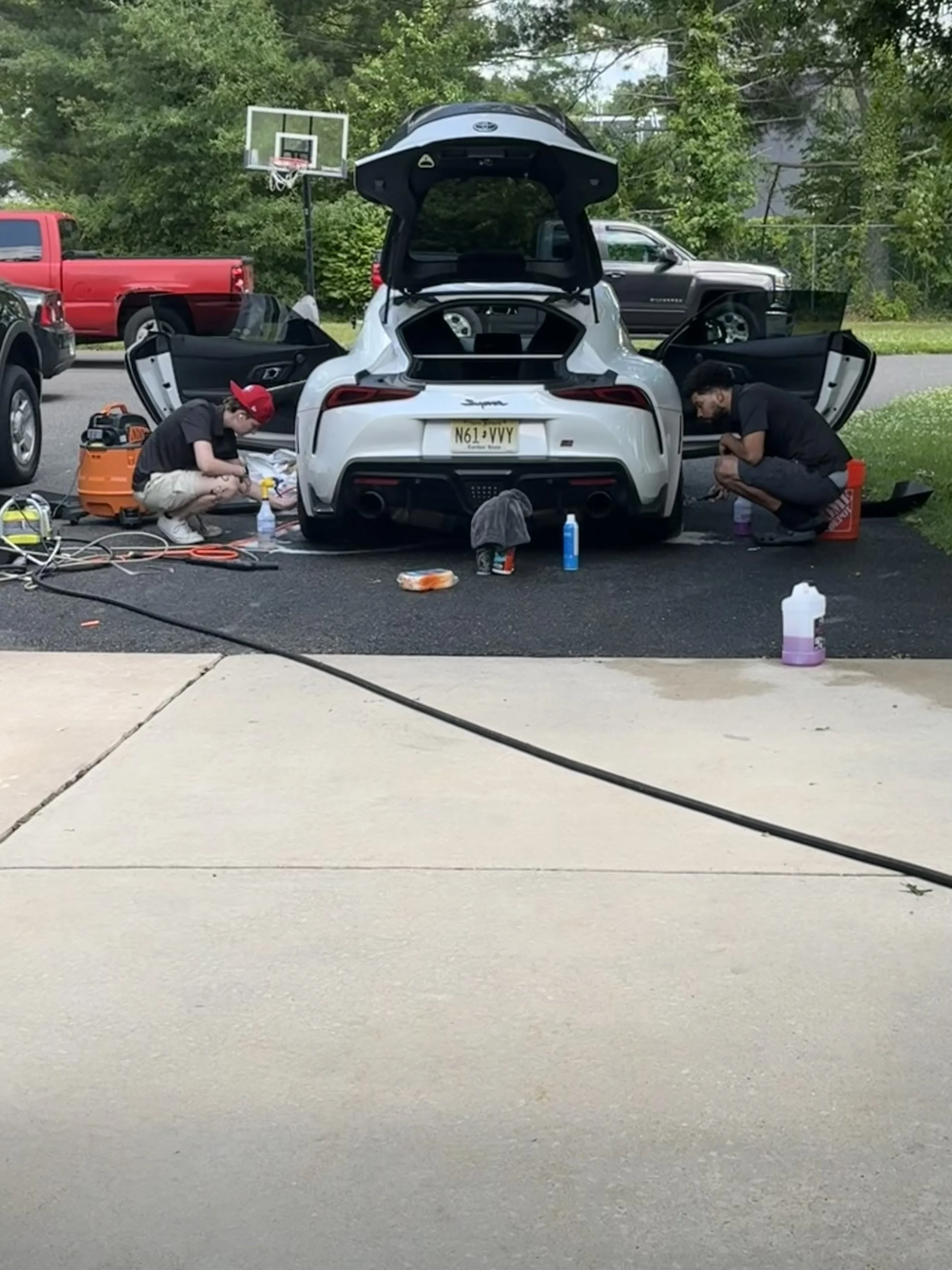 Two people cleaning a white sports car with the trunk and doors open, parked on a driveway.