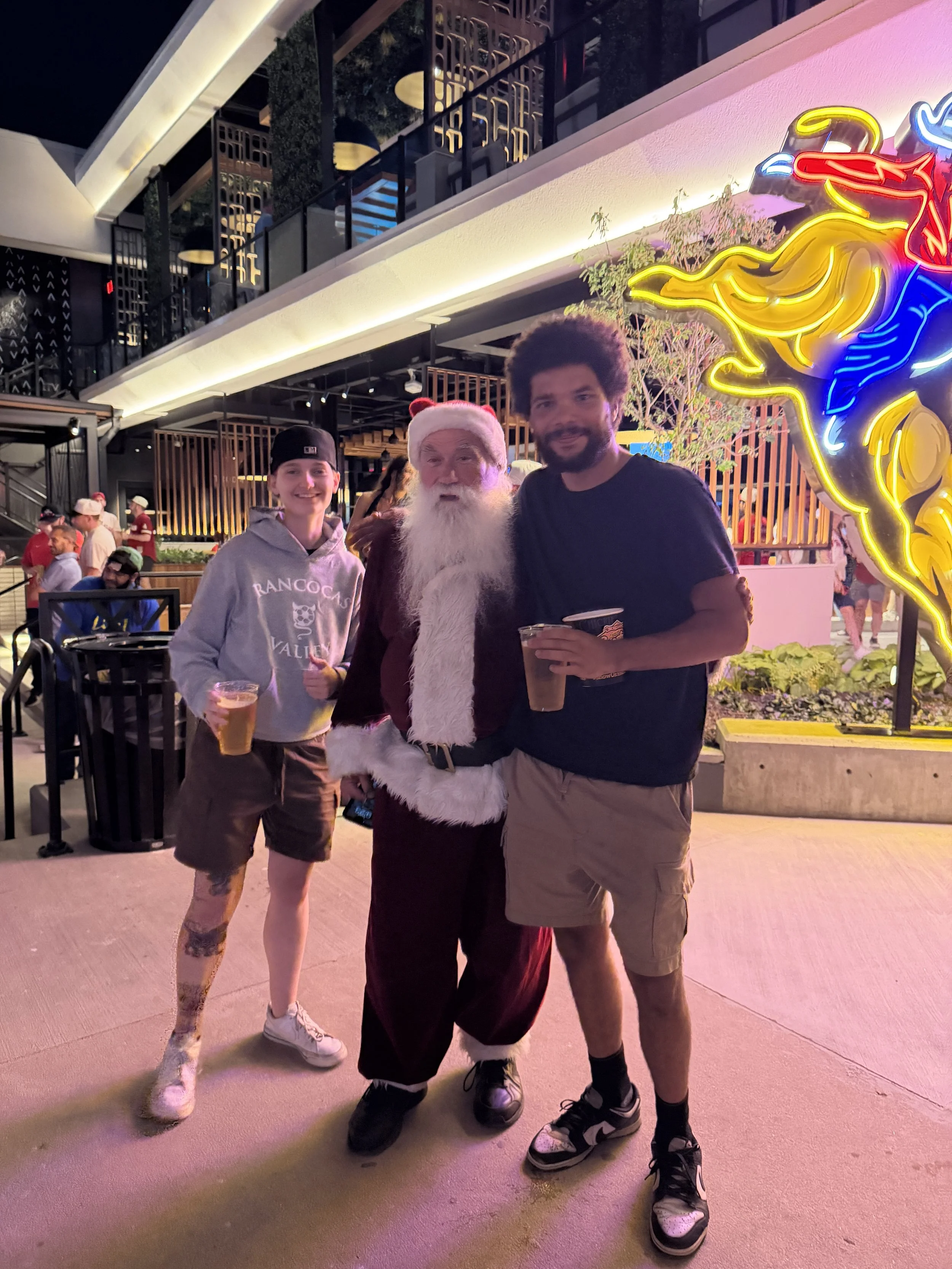 Three people standing together at night in an outdoor area with colorful neon lights, including a neon horse figure. The group includes a young man on the left wearing a gray hoodie and black cap, a person dressed as Santa Claus in the middle, and a man on the right with curly hair and a beard holding a drink. In the background, there are other people, tables, and a modern building with multiple levels.