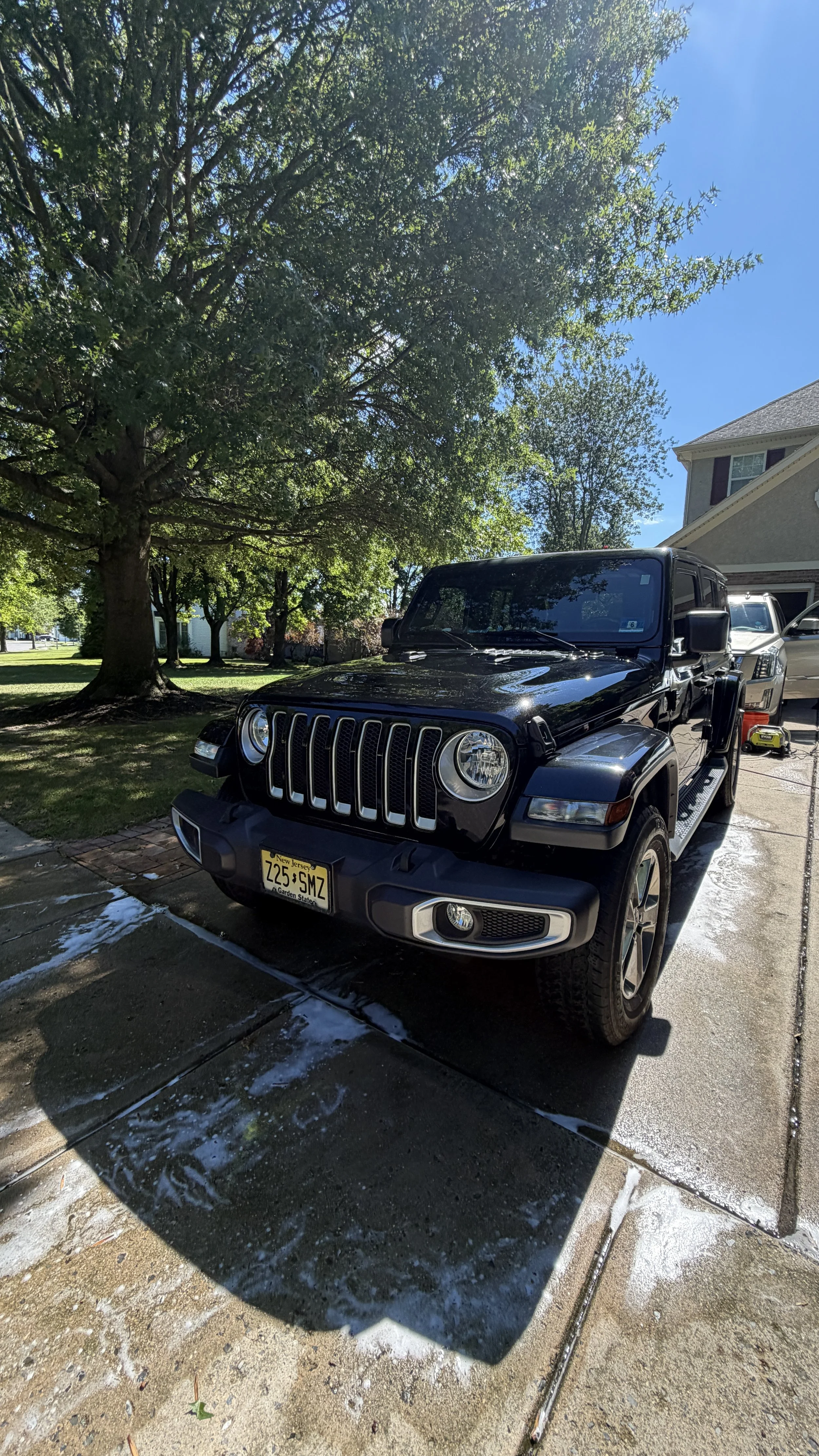 A black Jeep parked in a driveway with a tree and a house visible in the background on a sunny day. There is soap and water on the ground, indicating the vehicle was recently washed.
