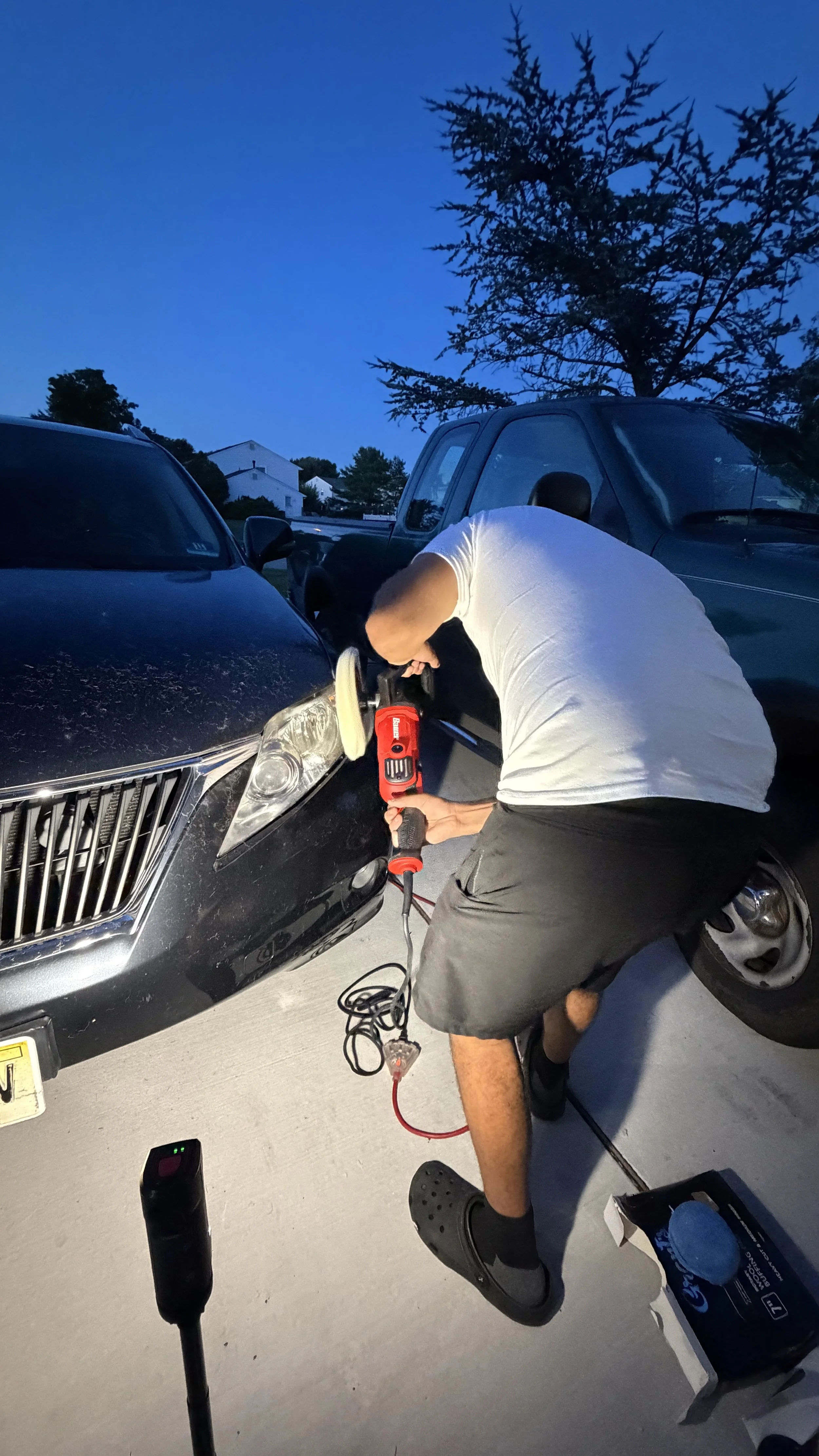 A man wearing a white t-shirt, black shorts, and Crocs is working on a black car with a power tool in a driveway during dusk. There are other parked vehicles and trees in the background.