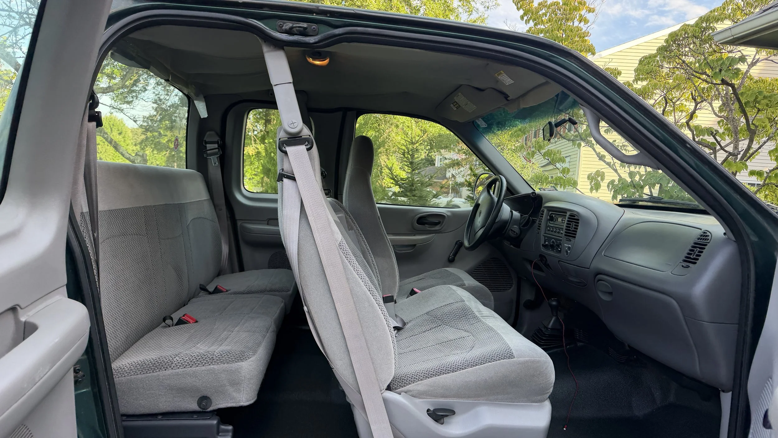 Interior of a pickup truck showing the front and back seats, dashboard, steering wheel, and seat belts, with a view of trees and a house outside.