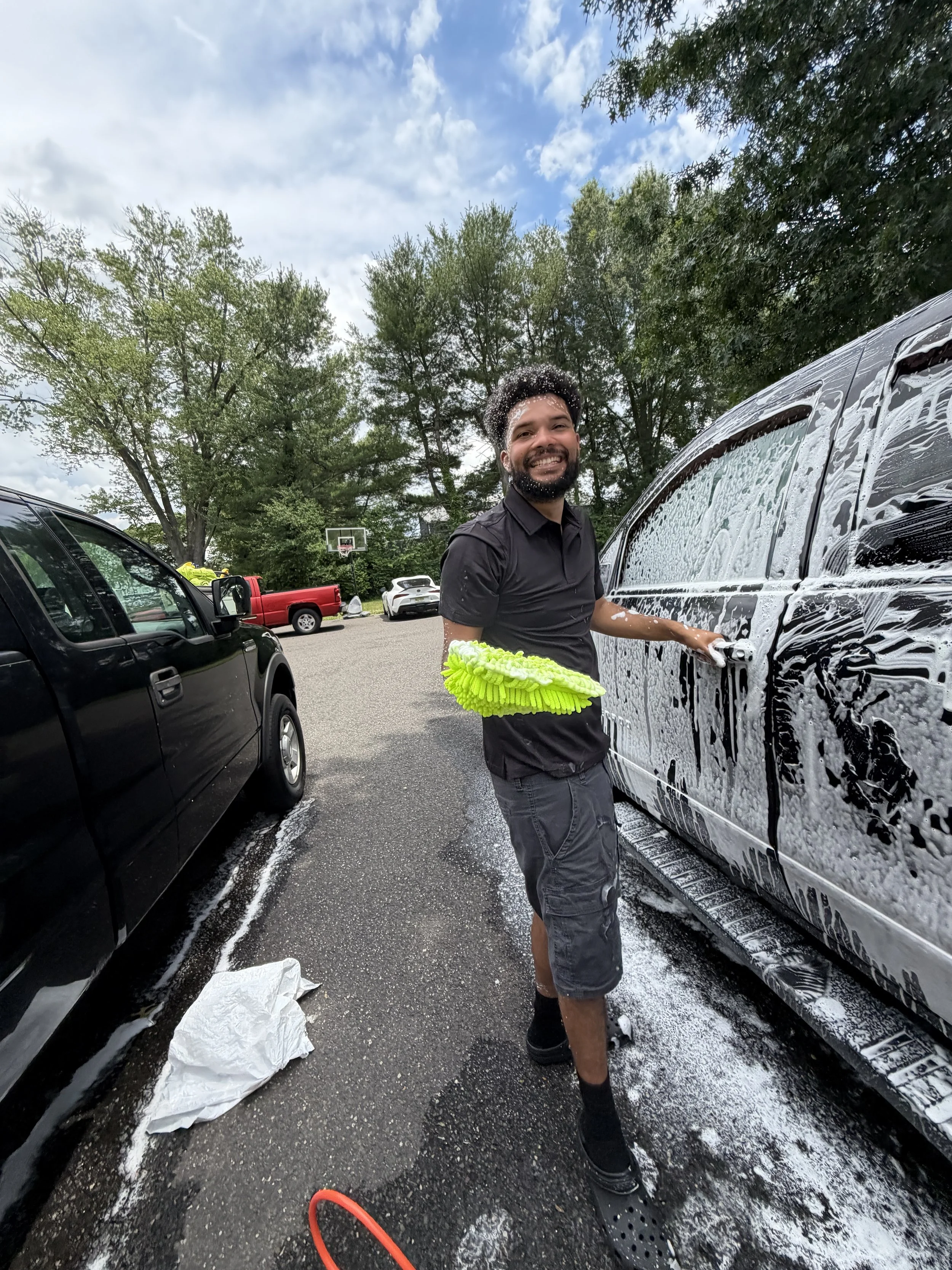 A man washing a black SUV with soap and a yellow sponge in a parking lot during the daytime. The man is smiling and standing next to the car, which is covered in soap suds. There are other cars parked nearby and trees in the background.