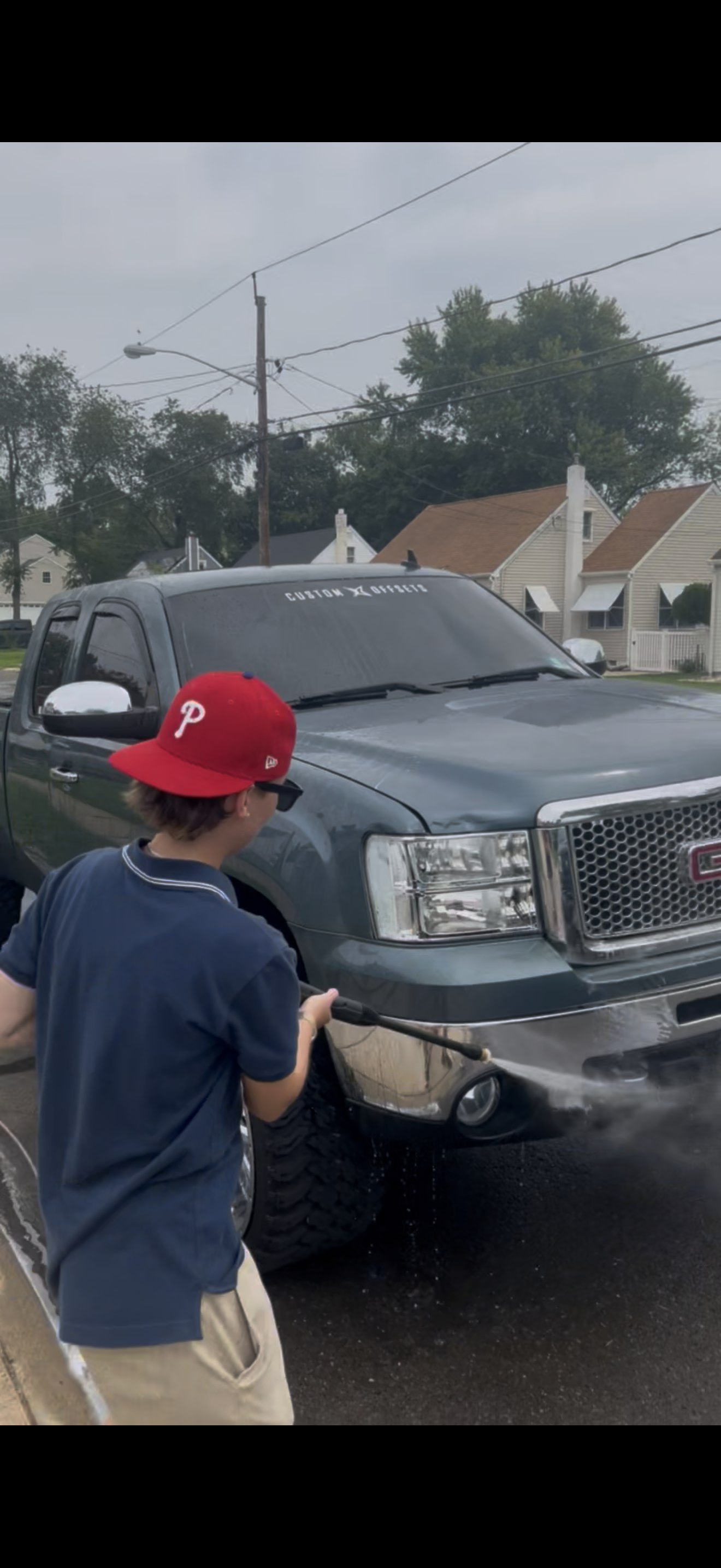 A boy wearing a red baseball cap, sunglasses, and a navy blue polo shirt, washing a gray GMC pickup truck in a parking lot. Houses and trees are in the background under an overcast sky.