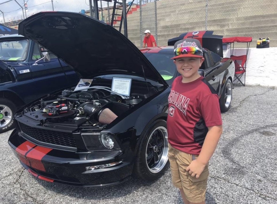 A boy smiling in front of a black sports car with its hood open at a car show. The boy is wearing a red South Carolina Gamecocks t-shirt, sunglasses, and a red cap, and standing on a paved area with other cars and a racing track in the background.