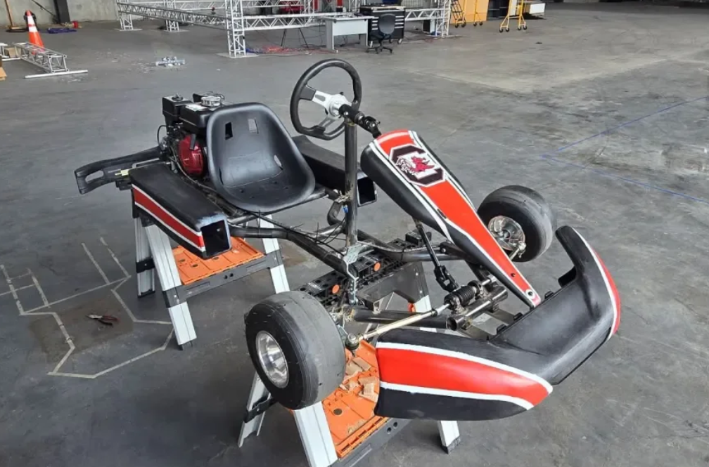 A go-kart chassis with a seat, steering wheel, and tires on a workshop floor, approximately mid-assembly or repair stage.