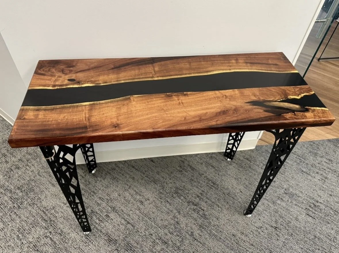 Wooden table with a black epoxy resin inlay running through the middle, supported by black metal lattice-style legs.