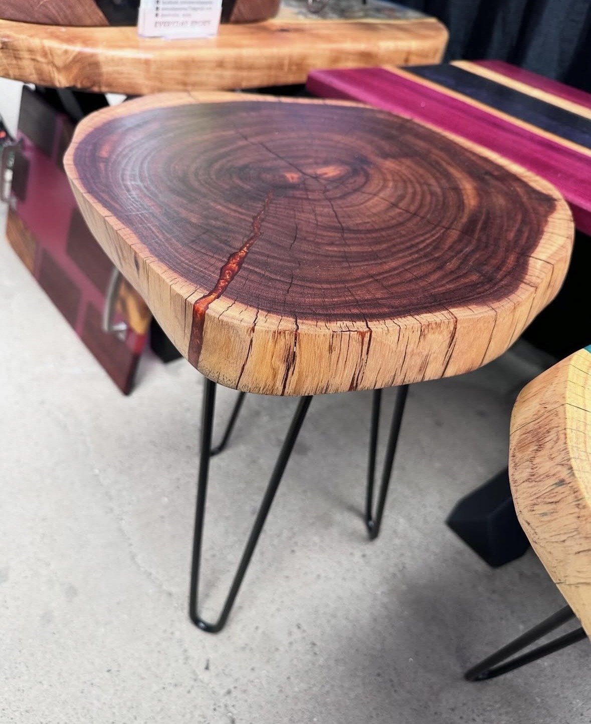 A wooden table with a natural edge and visible growth rings, enhanced with amber epoxy, supported by black metal hairpin legs, displayed at a store.