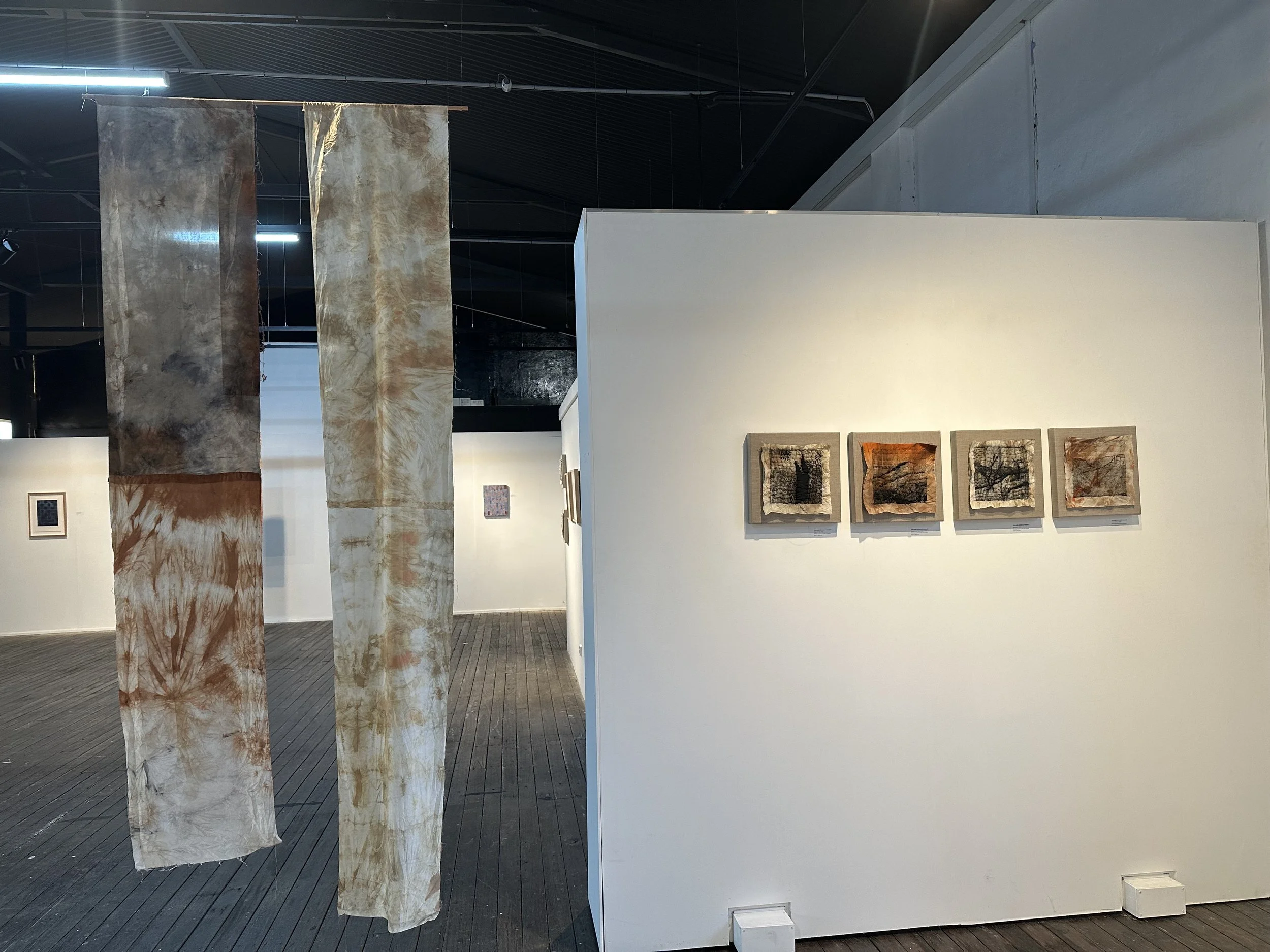 Art installation featuring hanging fabric pieces with earthy, tie-dye patterns beside a white wall showing framed textile artworks.