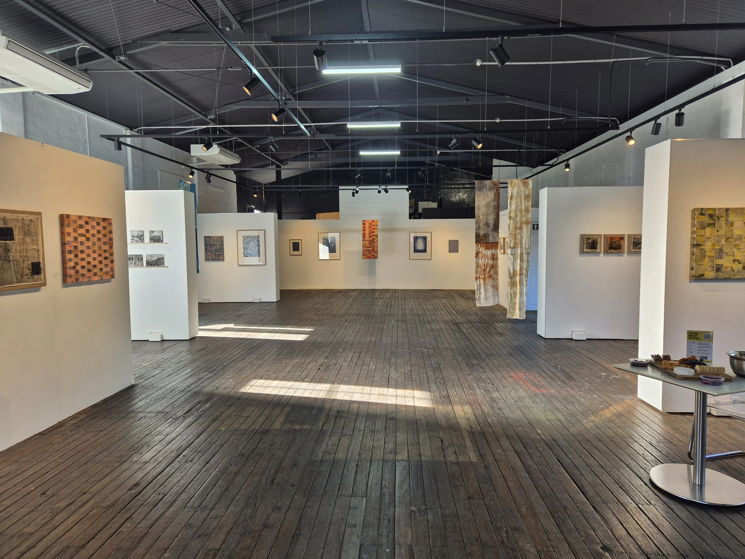 An art gallery with white walls displaying various paintings, with a dark ceiling featuring structural beams and track lighting, wooden flooring, and a few tables holding art supplies.