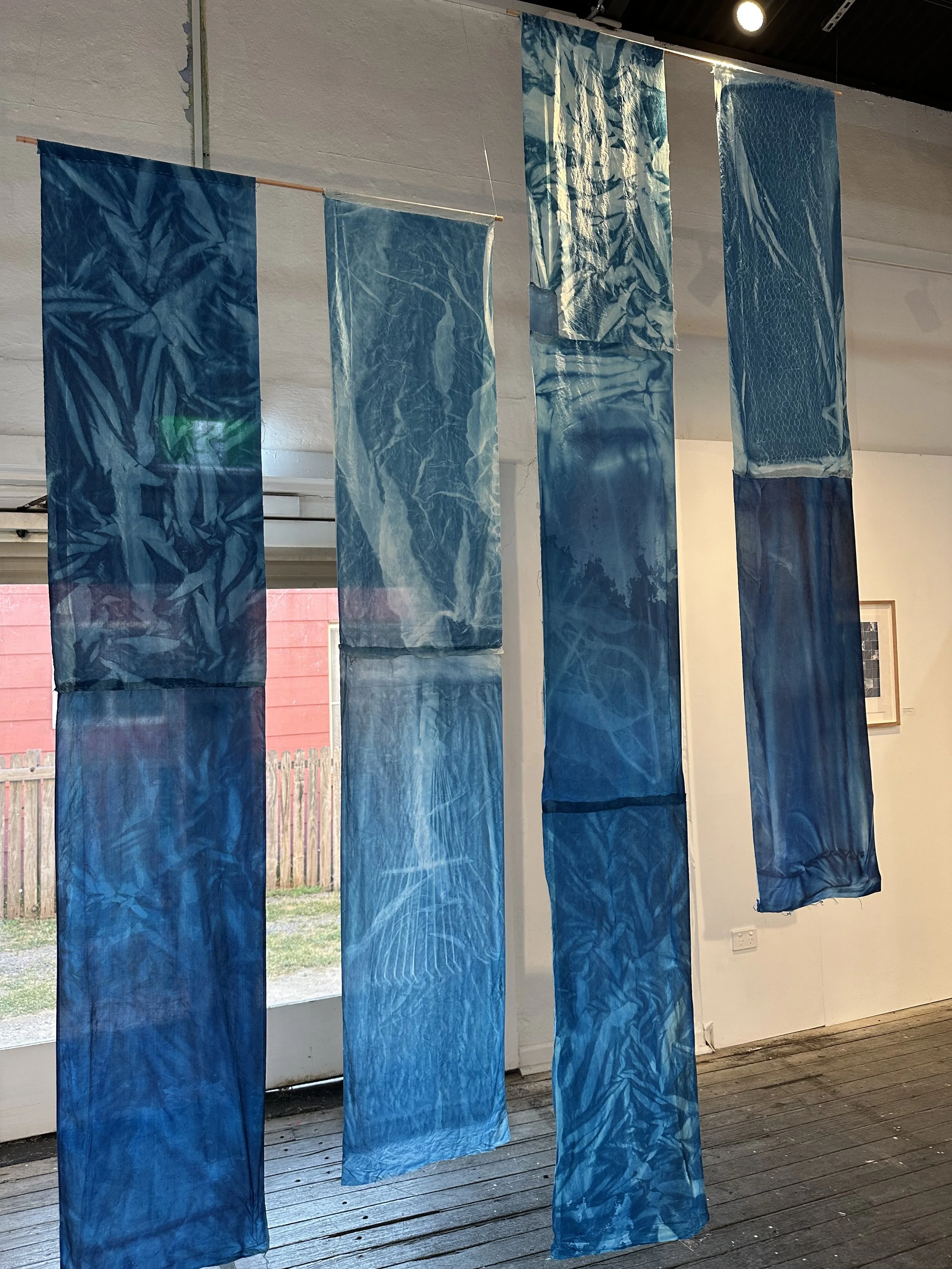 Five vertically hanging blue fabric pieces with different textures and patterns, displayed indoors near windows and a white wall.
