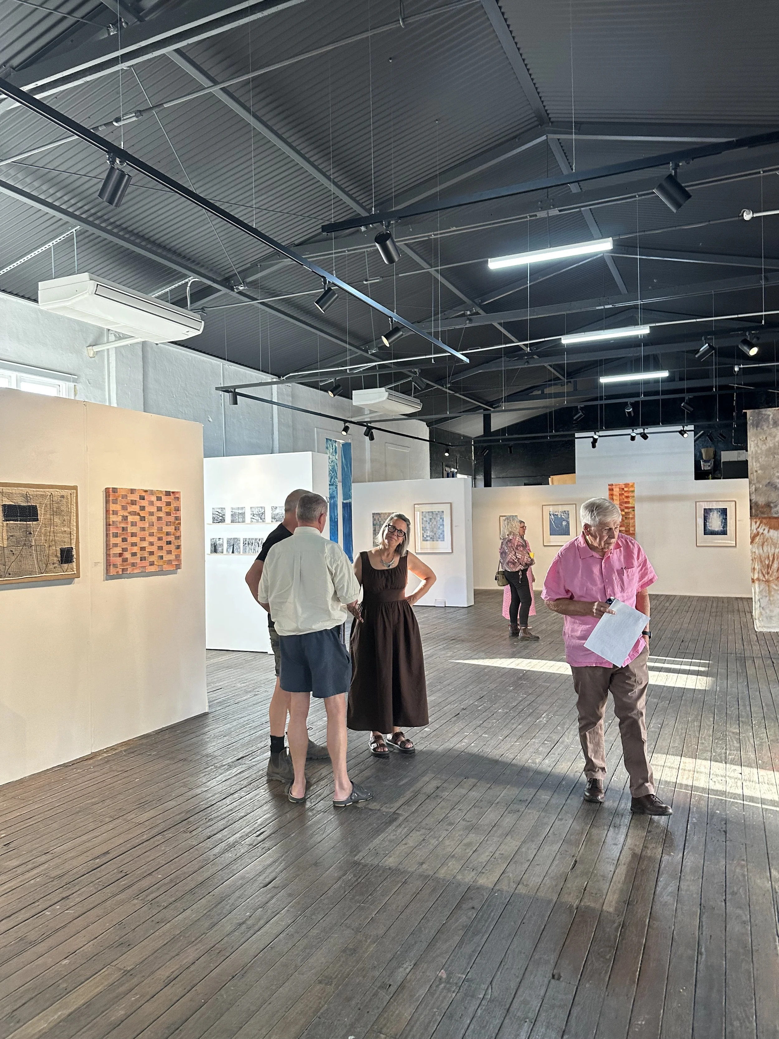 People viewing art pieces in an art gallery with wooden floors and white walls. Several artworks are displayed on the walls, and the ceiling has black metal beams and track lighting.