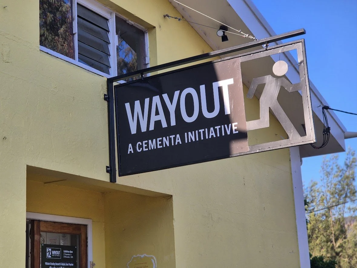 Sign for `WAYOUT`, a Cementa Initiative, hanging outside a yellow building with a window and a door, with trees and a clear blue sky in the background.