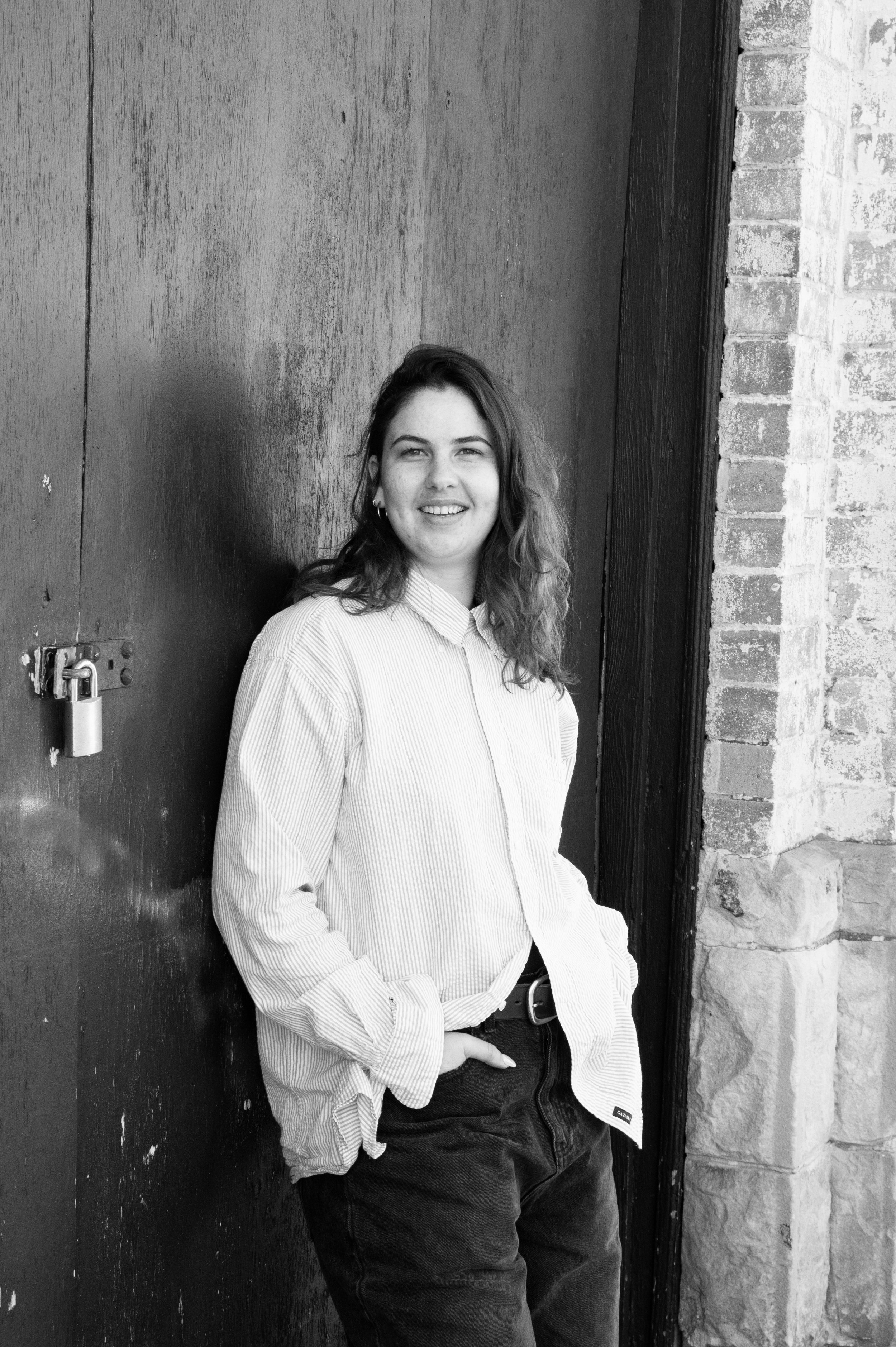 Black and white photo of a young woman with wavy hair, smiling, standing relaxed against a dark wooden door with a lock, in front of a brick wall.