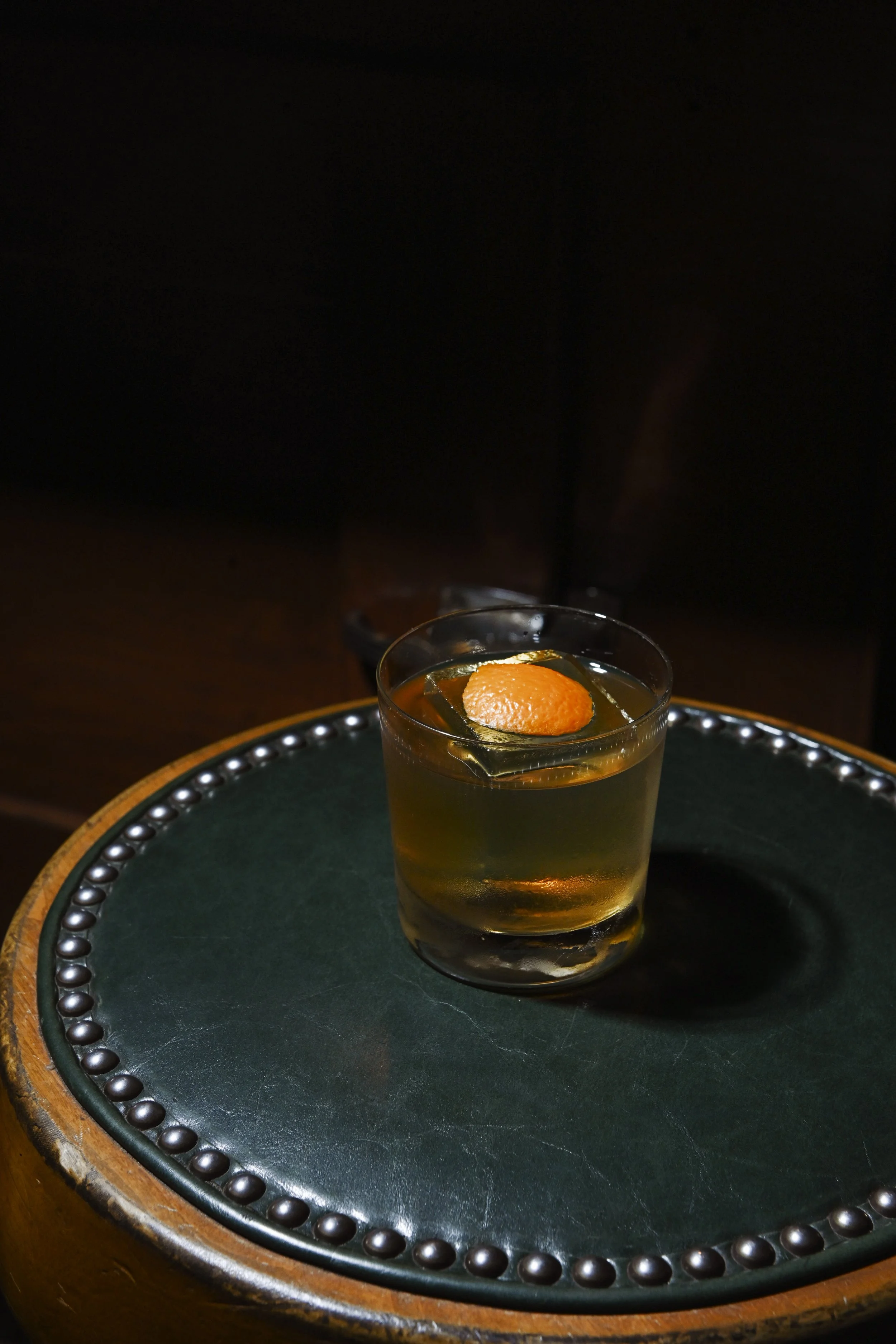 A glass of whiskey with an orange peel garnish on top, placed on a decorative tray with a green surface and a beaded border, against a dark background.