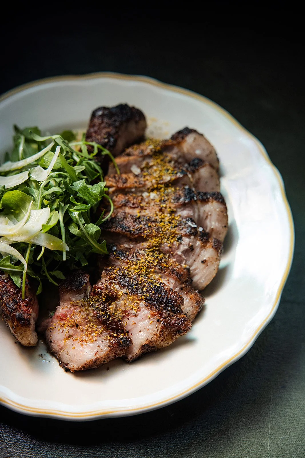 Grilled pork chop with seasoning, served with fresh greens on a decorative white plate.