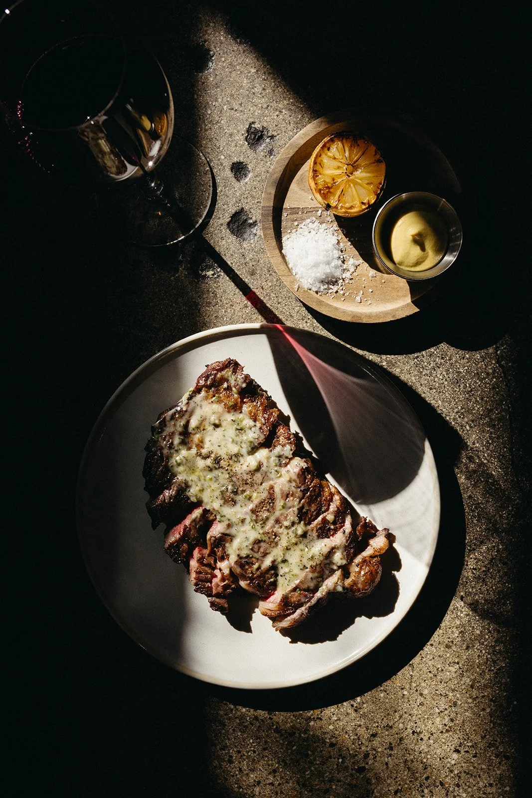 A plate of grilled steak, a glass of red wine, a halved grilled lemon, a small bowl of melted butter, and a small pile of salt, all on a rustic dark surface.