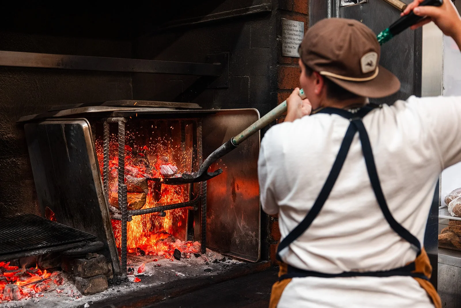 Person in a white shirt and brown hat tending a wood fire in a brick oven.