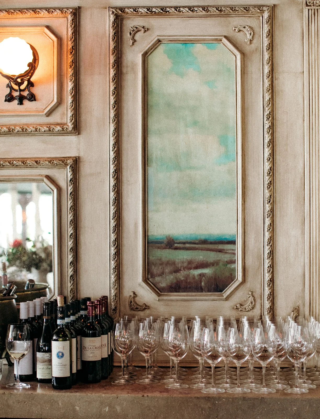 Interior with wine bottles and glasses on a counter, a large landscape painting, and ornate wall details.