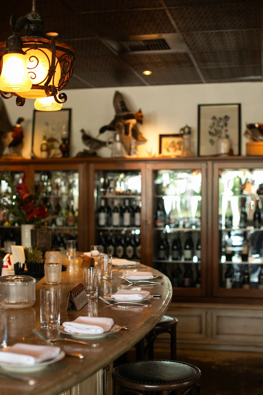 A cozy restaurant interior featuring a wooden bar counter set with glasses, napkins, and utensils, with wine bottles in a glass-fronted cabinet behind the bar, decorative artwork and duck sculptures on the wall above, and a warm, ambient lighting fix