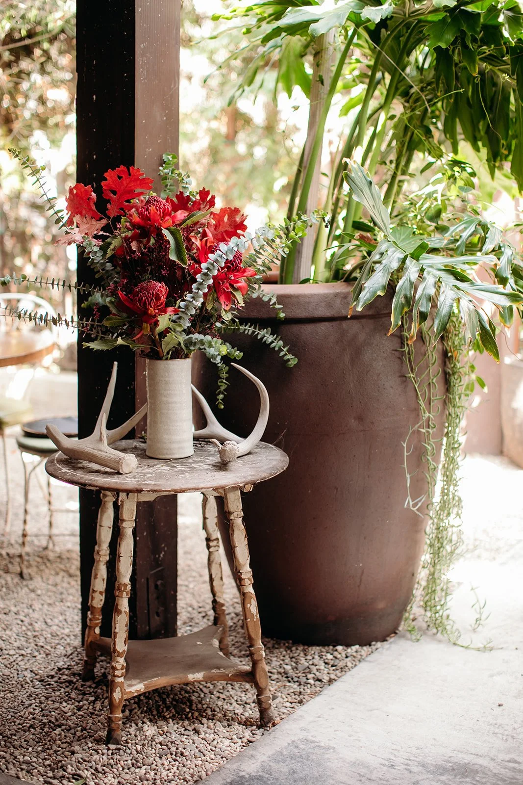 A small wooden stool holding a beige vase with red and green floral arrangement, a large potted plant with green leaves, and decorative antlers.