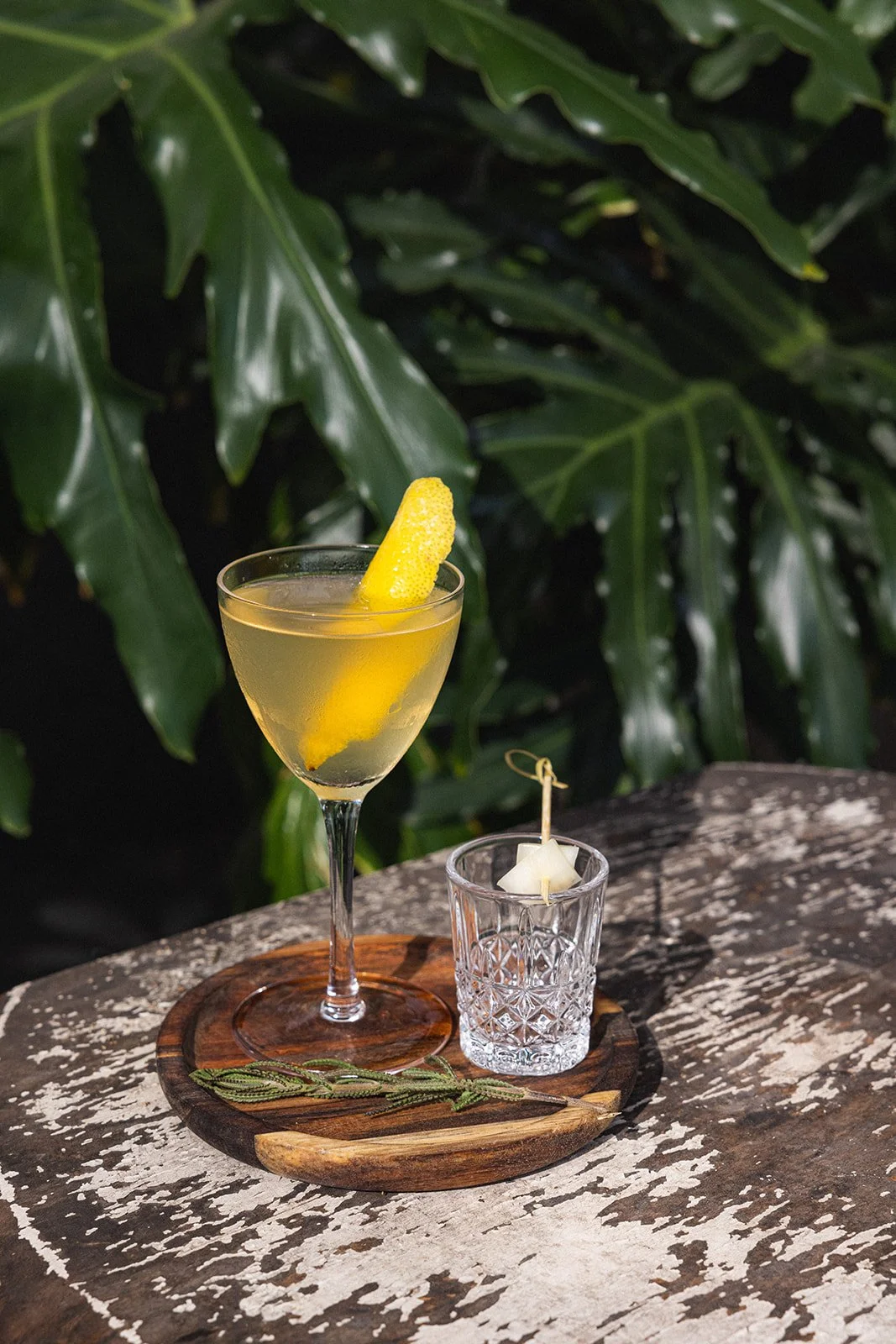 A cocktail glass with a lemon twist garnish, placed on a wooden tray with a sprig of herbs, on a weathered wooden surface with green leaves in the background.