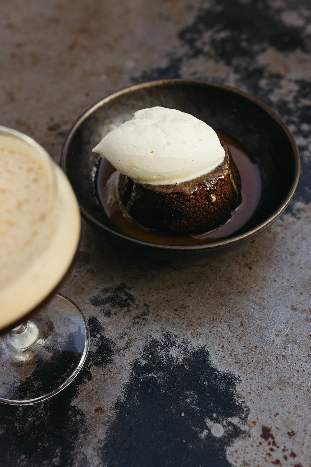 A date cake topped with vanilla ice cream in a small black bowl on a rustic surface.