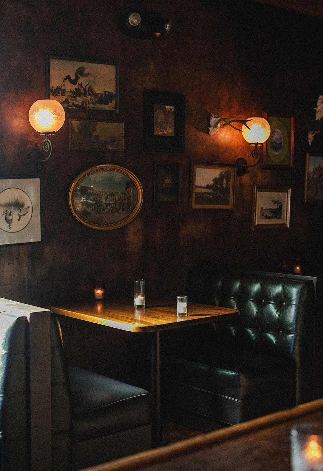 A dimly lit cozy restaurant corner with dark wood walls, framed art, vintage light fixtures, a dark leather booth with glasses of water on the table.