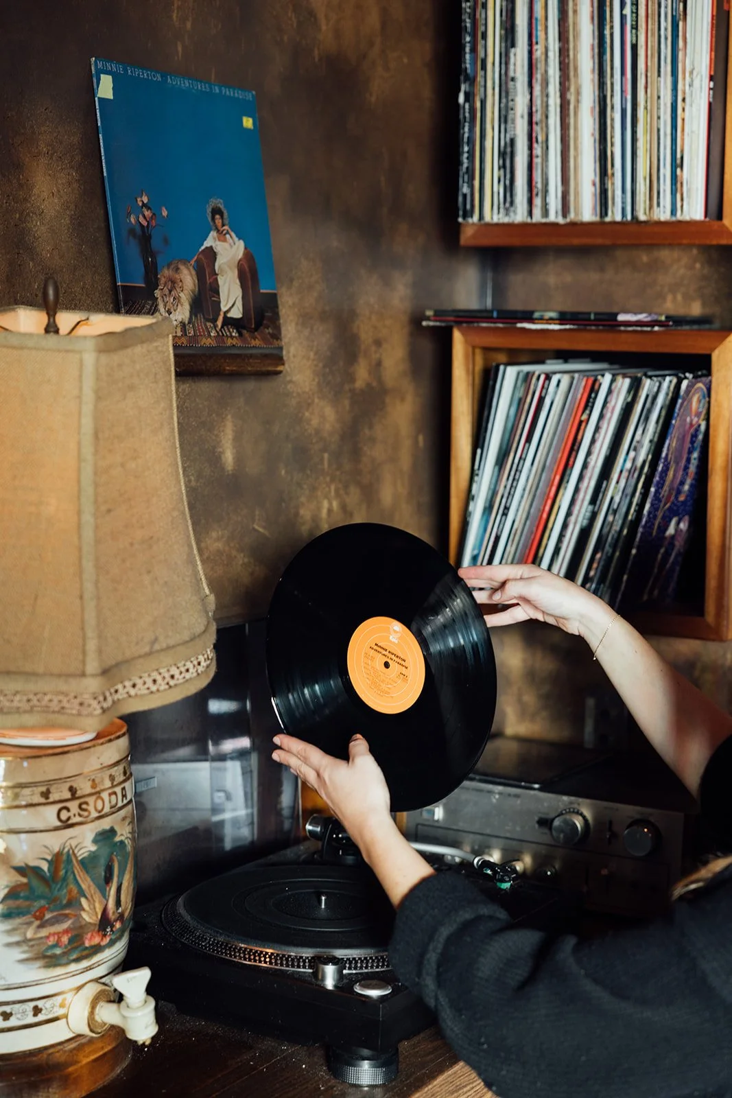Person placing a vinyl record on a turntable in a room with a bookshelf filled with records and a vintage lamp.