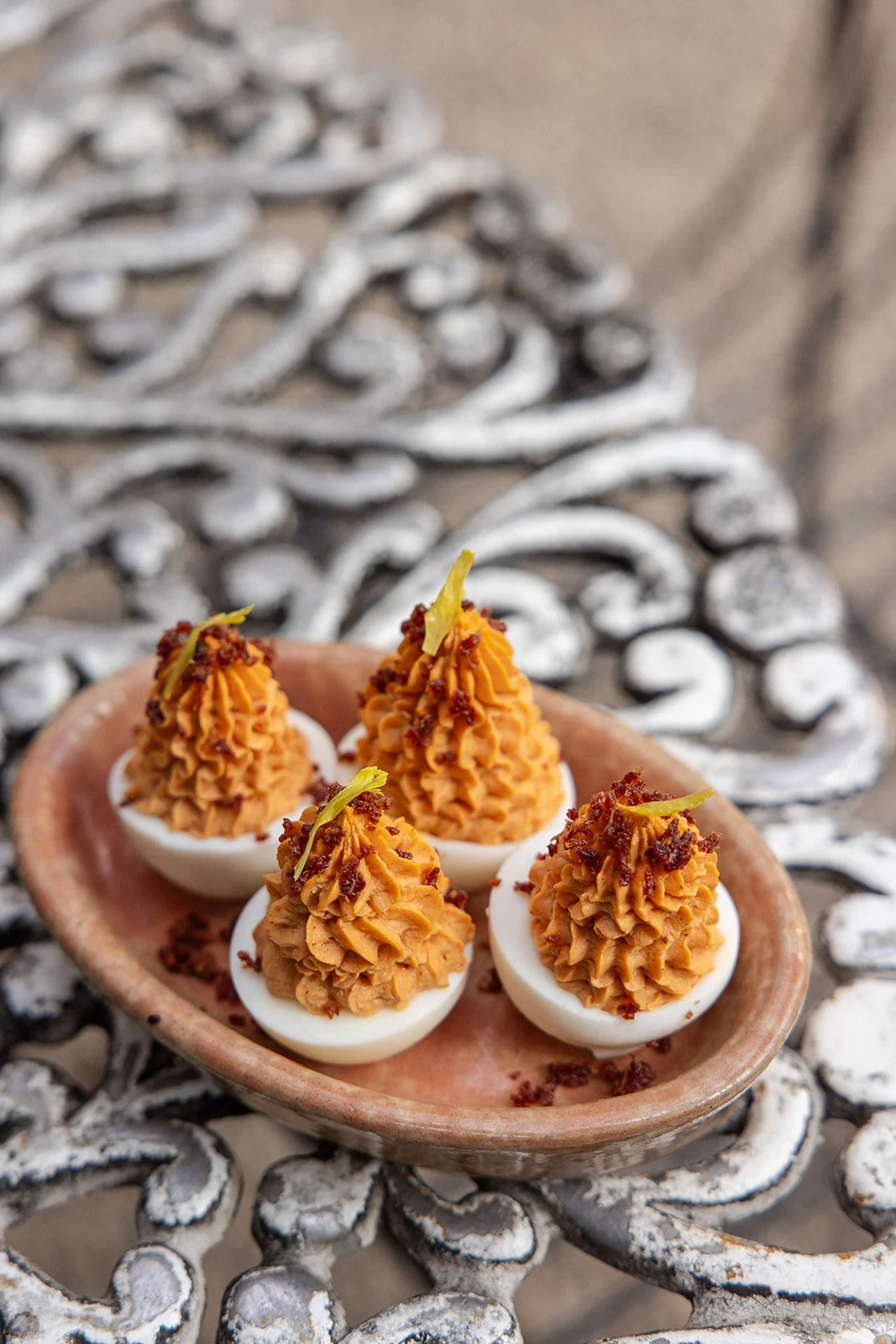 Four stuffed eggs topped with piped orange-colored filling and garnished with small yellow-green herbs on a ceramic bowl, placed on an ornate metal table.