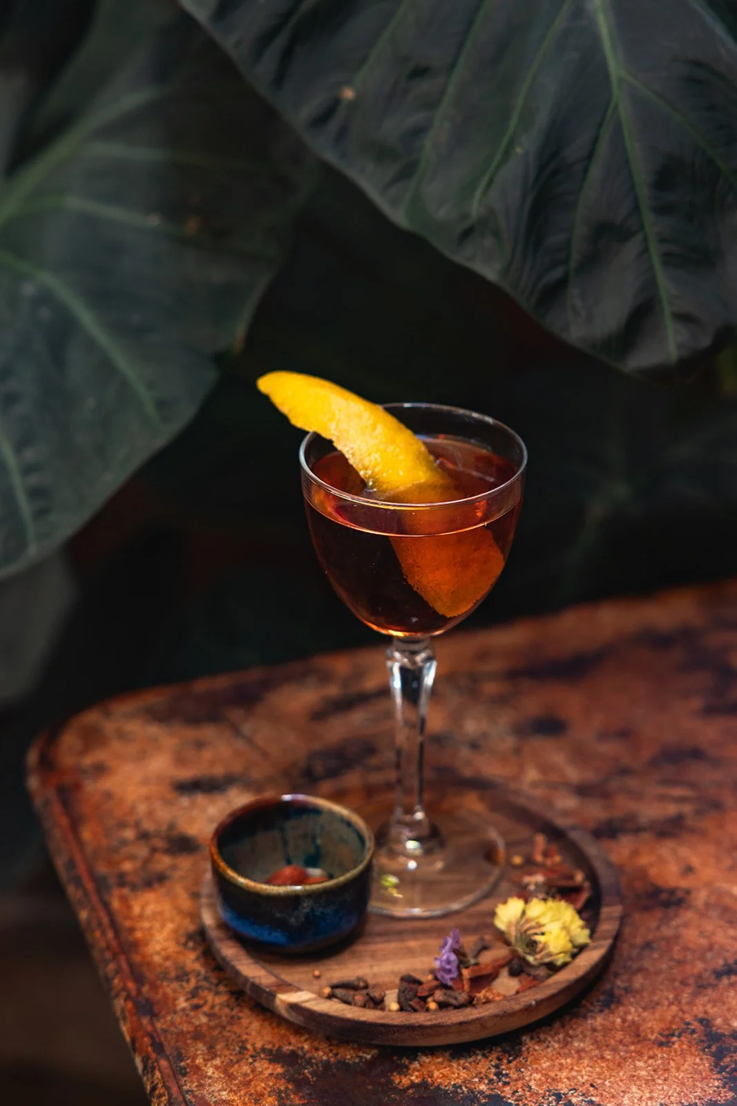 A glass of dark red cocktail with a lemon wedge garnish on a wooden table, accompanied by a small bowl and decorative herbs and flowers.