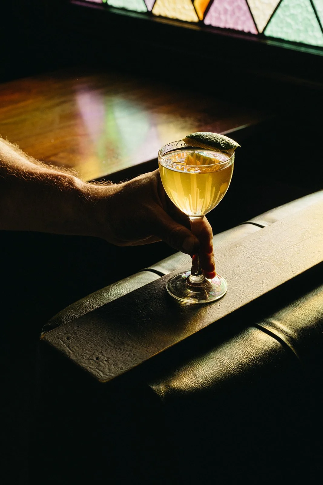 A person holding a cocktail with lemon garnish in front of a stained glass window, casting colorful light on a dark surface.