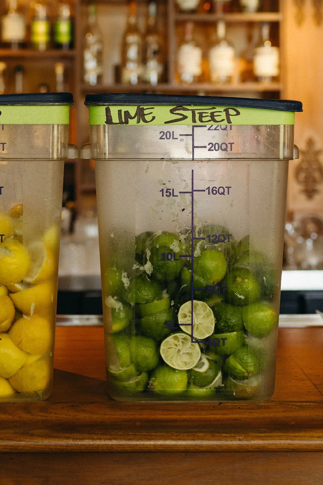 A plastic container filled with halved limes and lime slices, labeled 'LIME STEEP' on the top edge, placed on a wooden surface in a bar or kitchen setting.