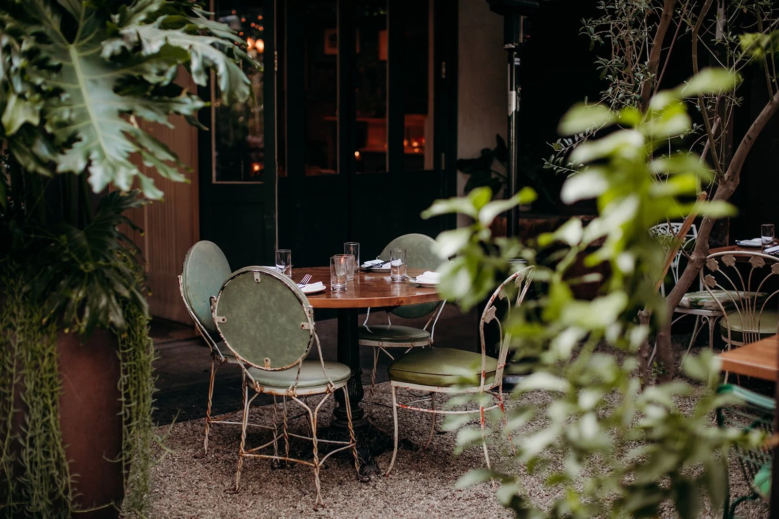 Outdoor dining area with a round wooden table set with glasses and utensils, surrounded by vintage green chairs, and lush green plants and trees.