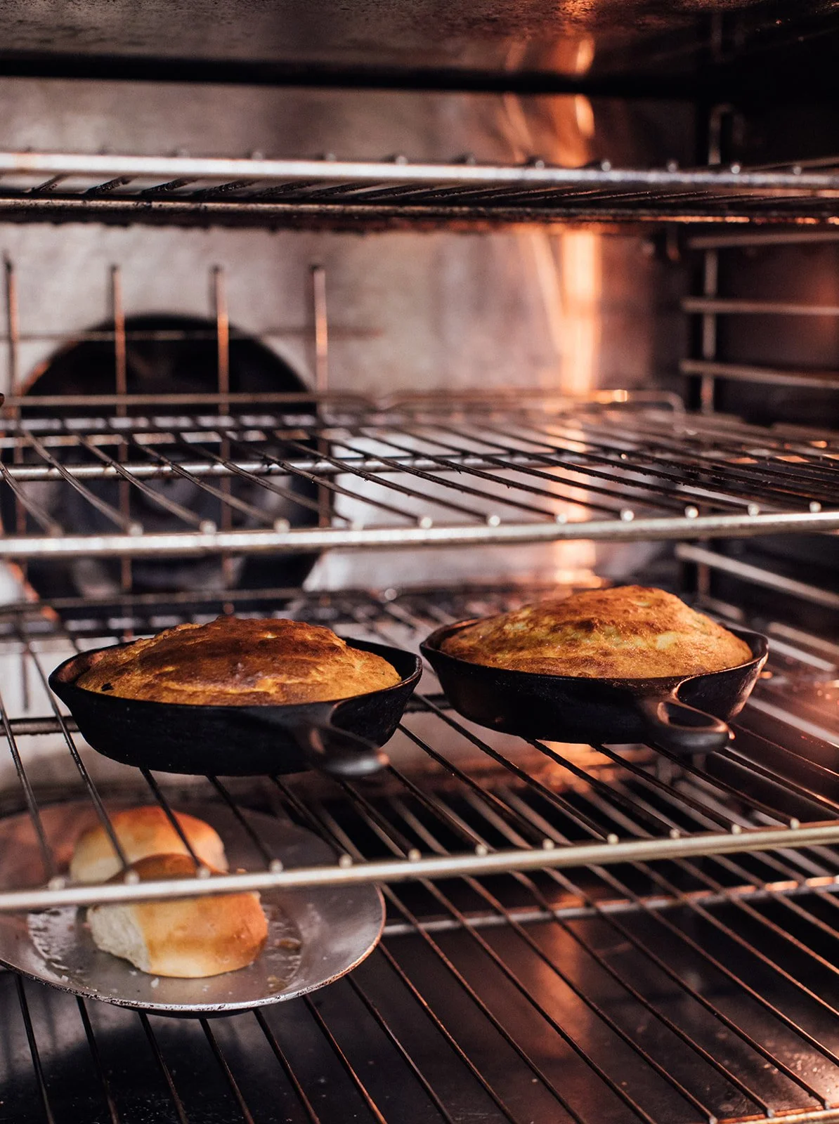 Two bowls of freshly baked bread in a kitchen oven with racks and a piece of bread on a plate in the foreground.