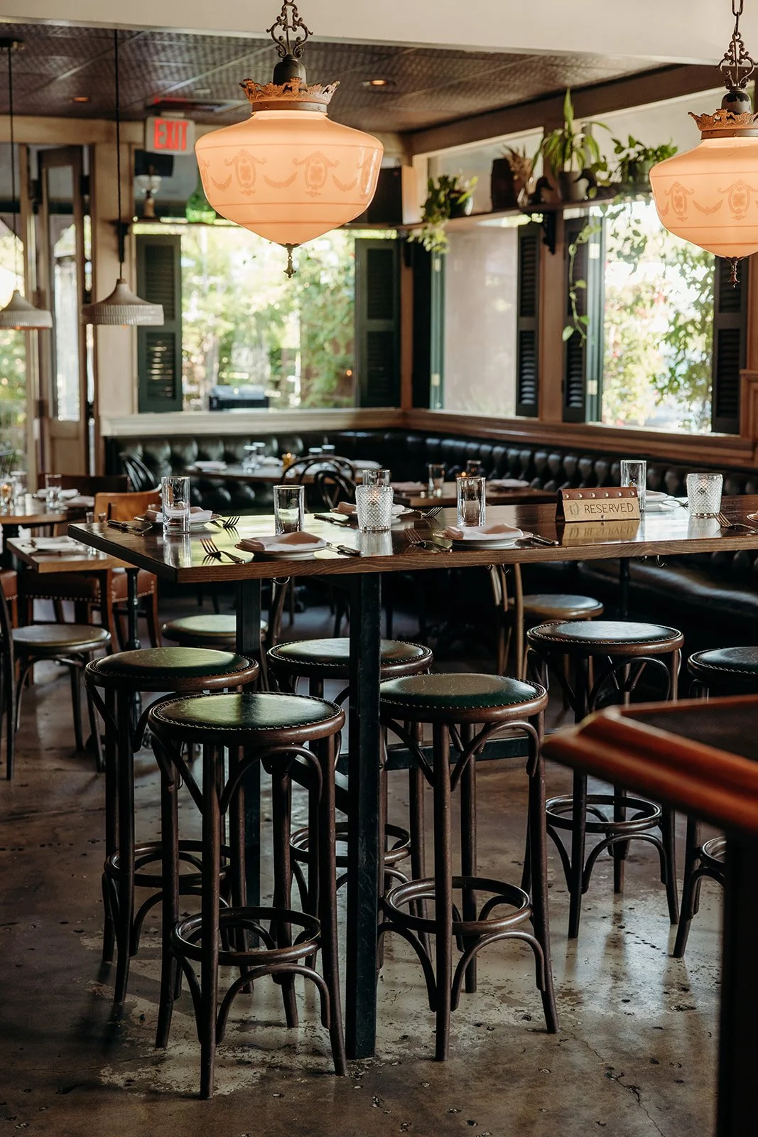 Interior of a cozy restaurant with wooden tables set for dining, including glasses, napkins, and cutlery. Black leather banquette seating along the walls, large windows with green shutters letting in natural light, and pendant and chandelier lighting