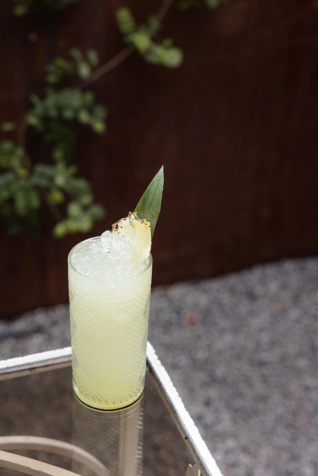 A light yellow cocktail with ice in a tall glass, garnished with a pineapple slice and a pineapple leaf, on a metal surface outdoors.