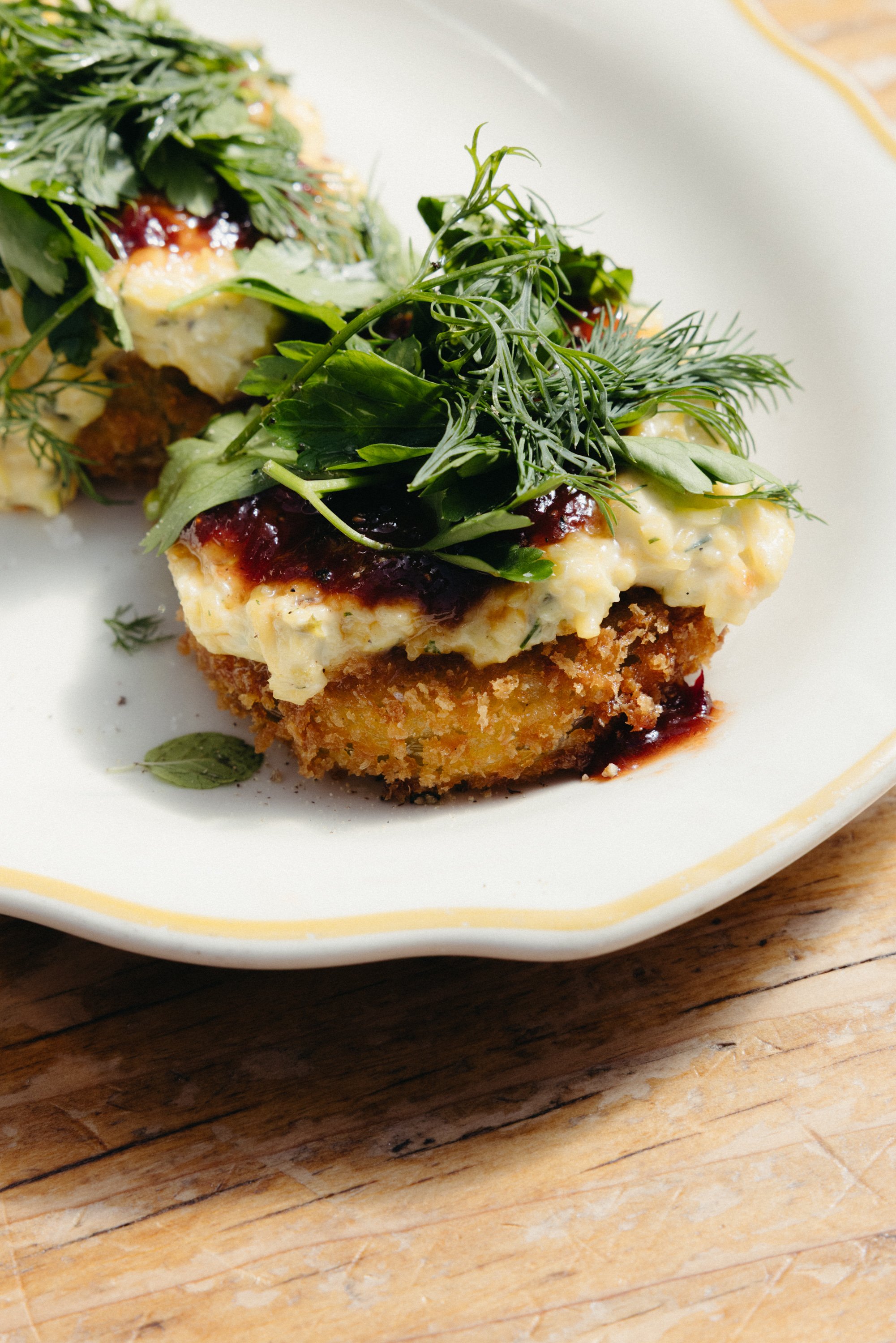 Close-up of a breaded fried chicken topped with mashed potatoes, gravy, and fresh herbs on a white plate, sitting on a wooden surface.