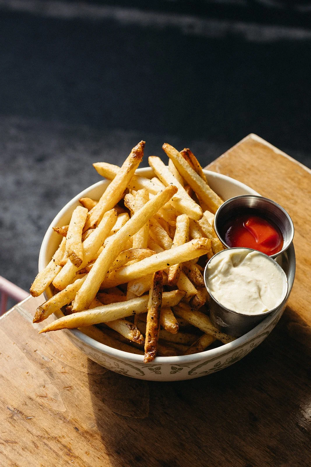 A bowl of French fries with two small metal containers of ketchup and mayonnaise on a wooden surface.