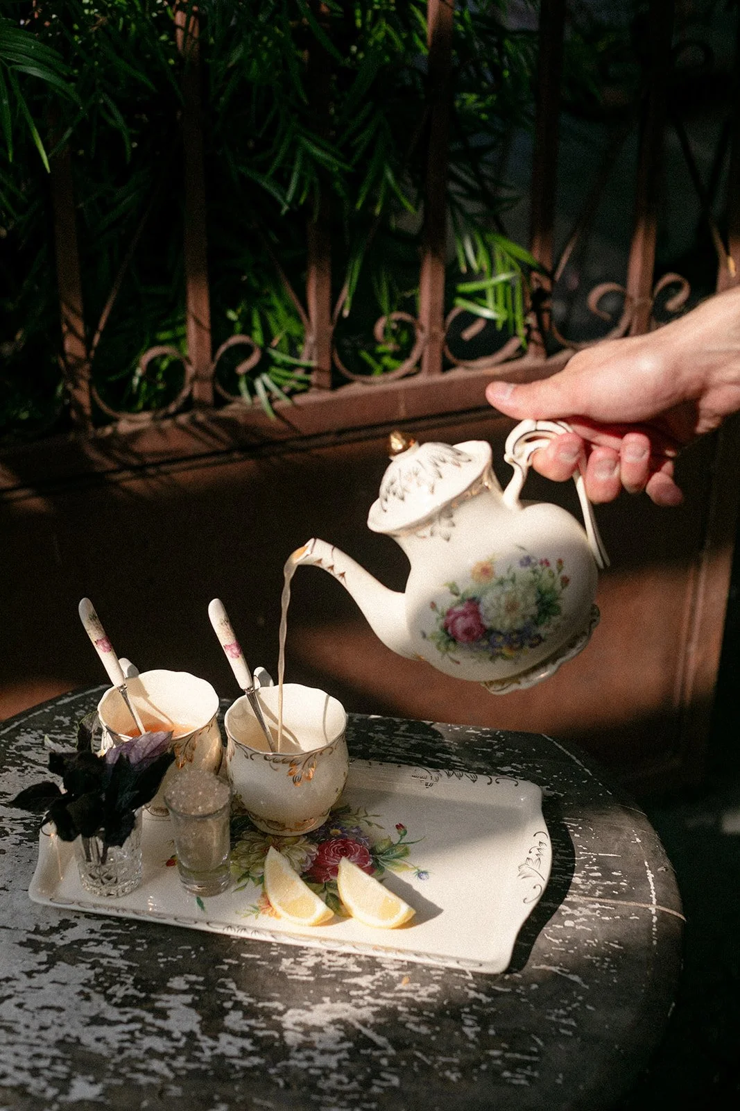 A person pouring tea from a floral teapot into a cup on a decorative tray with lemon slices and small cups, set on a worn wooden table with a dark, ornate background.