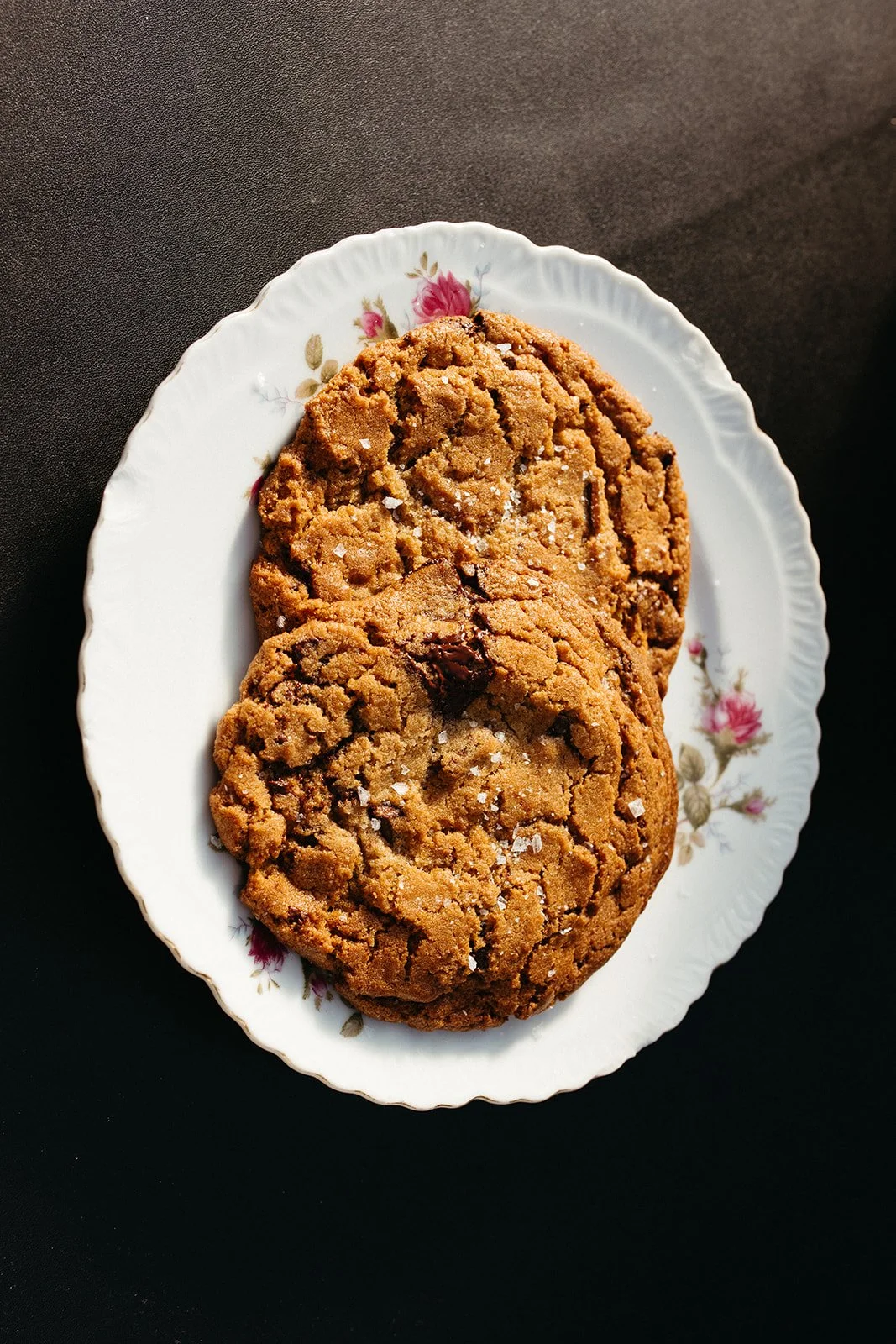 Two chocolate chip cookies on a floral-patterned white plate with scalloped edges, placed on a dark surface.